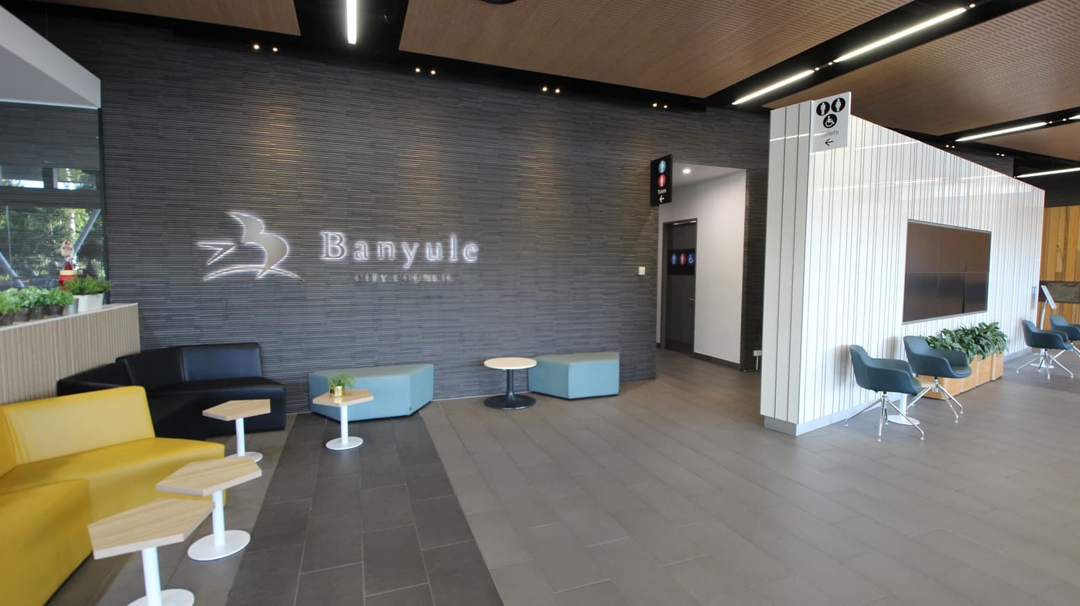 A modern lobby area with a grey tiled floor and a wooden panelled ceiling. On the left, there are yellow and black sofas with small hexagonal tables. The wall features the word "Banyule" with a bird logo. On the right, a white panelled wall has a mounted screen and blue chairs with a planter beneath. A corridor leads to restrooms, indicated by signs above. A window on the far left shows greenery outside.