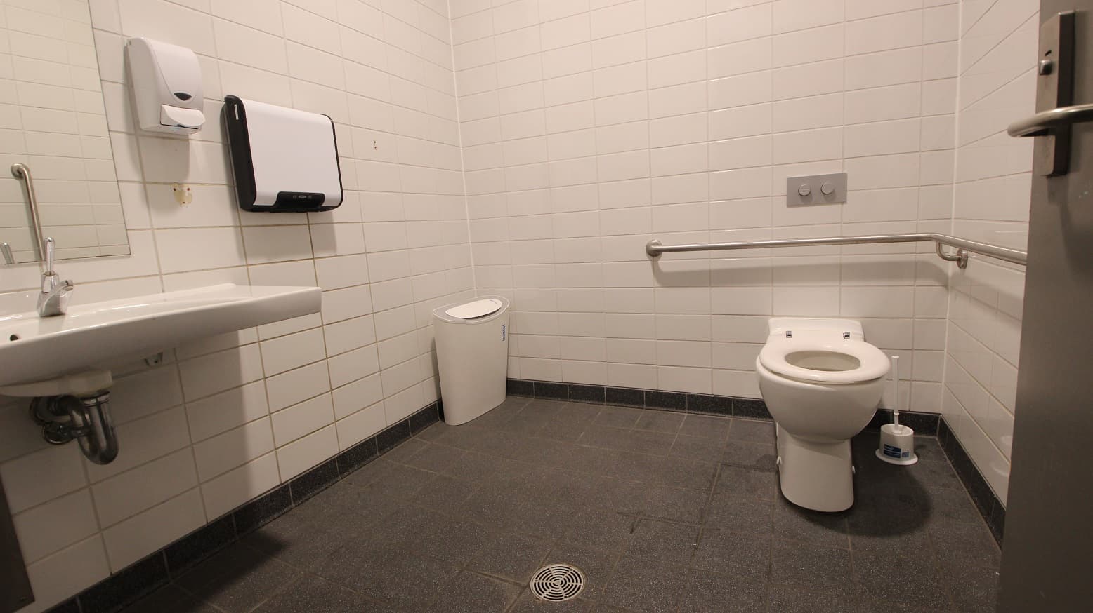 A public restroom with white tiled walls and a grey tiled floor. On the left, a wall-mounted sink with a soap dispenser and a paper towel holder above it. In the middle, a white waste bin is placed against the wall. On the right, a toilet with a grab bar beside it and a toilet brush on the floor. A metal door handle is visible on the right edge.
