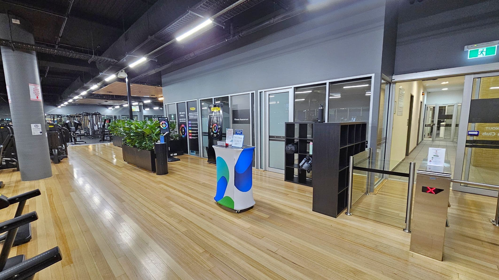 A gym interior with a wooden floor and a dark ceiling. On the left, there are exercise machines and a large grey pillar. In the centre, a small table with a colourful cover is placed near a row of glass-walled rooms labelled for fitness assessments. A black shelving unit is nearby. On the right, a turnstile with a red 'X' and a glass door leading to a hallway. Bright overhead lights illuminate the space, and a green exit sign is visible above the door.