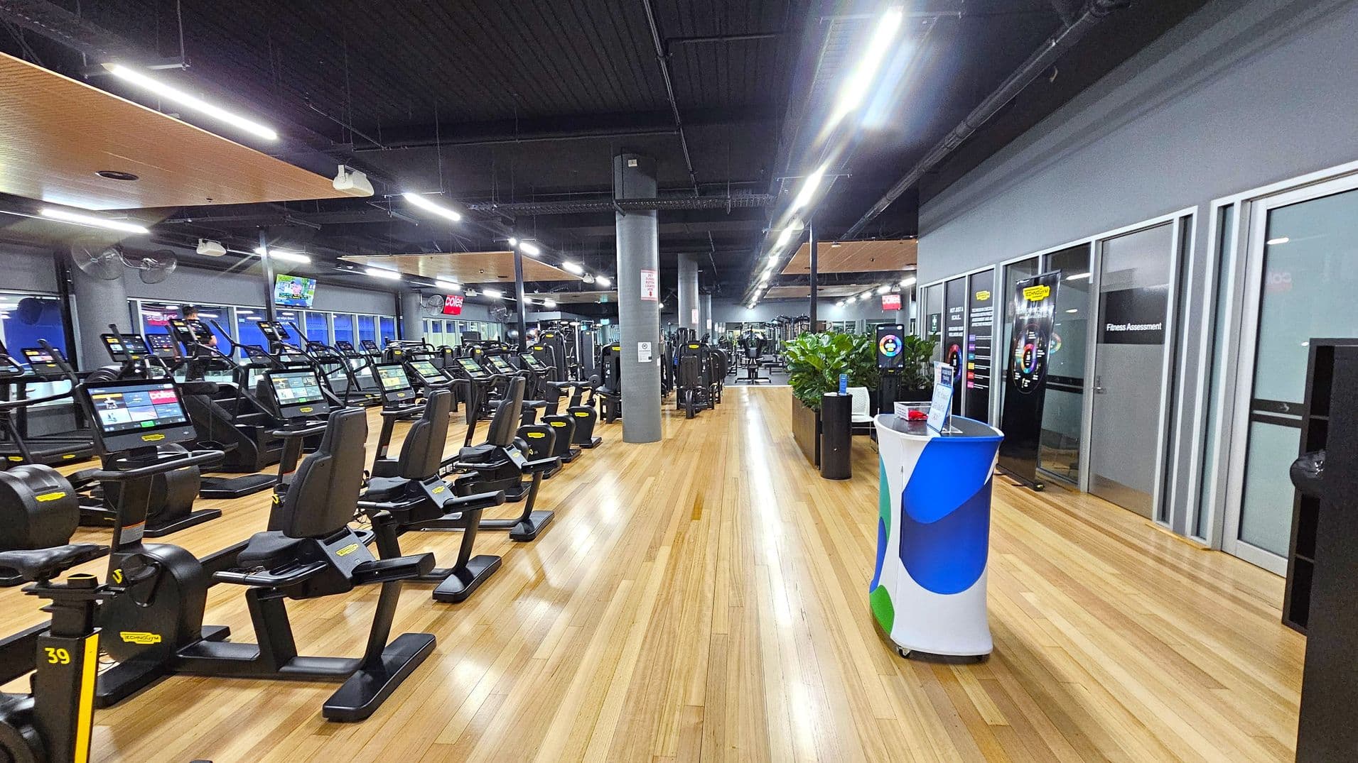 A spacious gym interior with a wooden floor and a dark ceiling. On the left, there are rows of exercise machines, including treadmills and stationary bikes, each equipped with digital screens. In the middle, a wide walkway leads to more equipment in the background. On the right, there are glass doors labelled "Fitness Assessment" and a small counter with a colourful cover. Potted plants are placed near the counter, adding greenery to the space. Bright overhead lights illuminate the area.