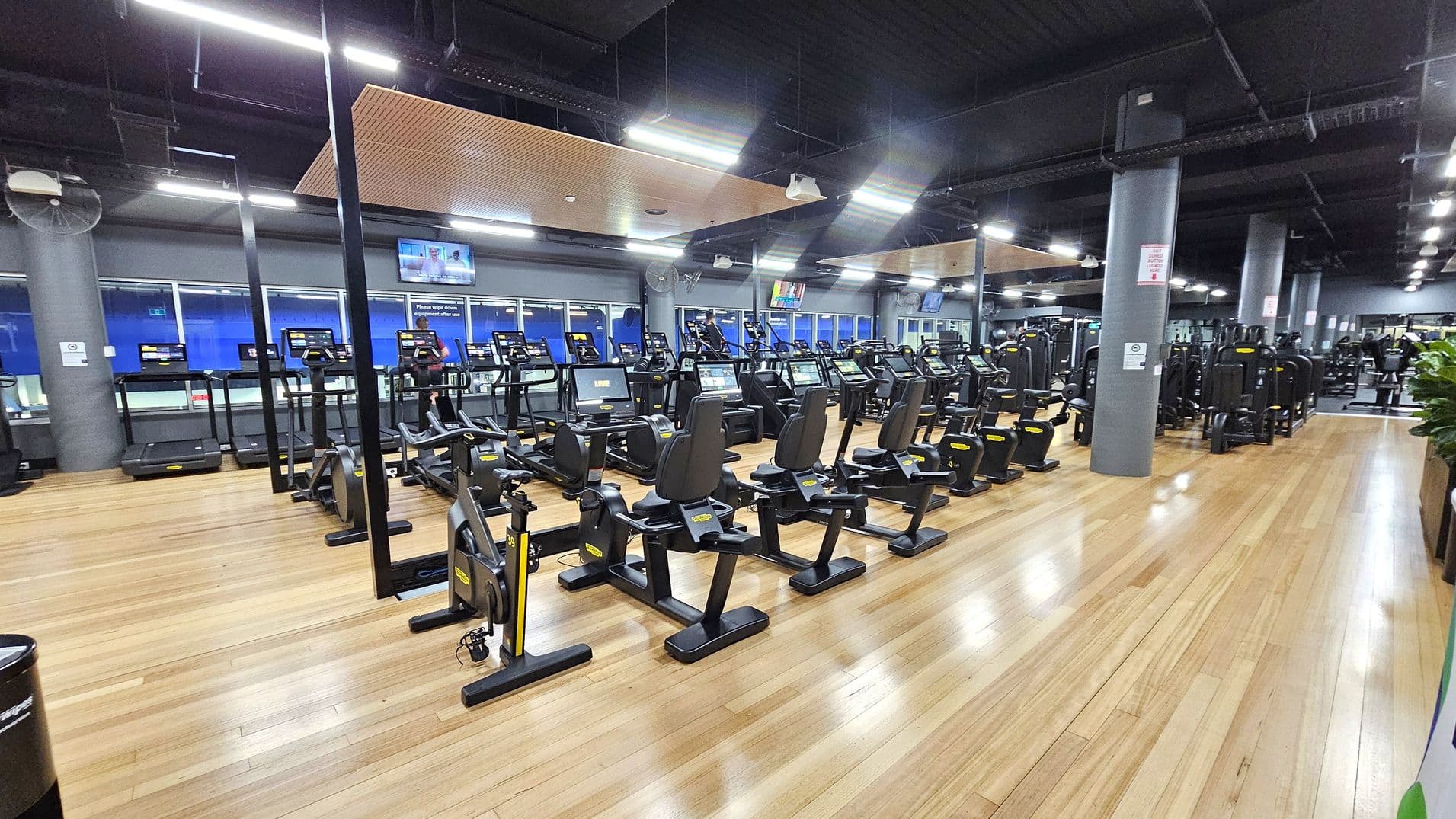 A spacious gym with wooden flooring and a dark ceiling. In the foreground, there are several stationary bikes and treadmills arranged in rows. The background features large windows and multiple television screens mounted on the walls. The gym is well-lit with overhead lights, and there are grey pillars throughout the space. On the right, there are additional exercise machines and a row of plants.