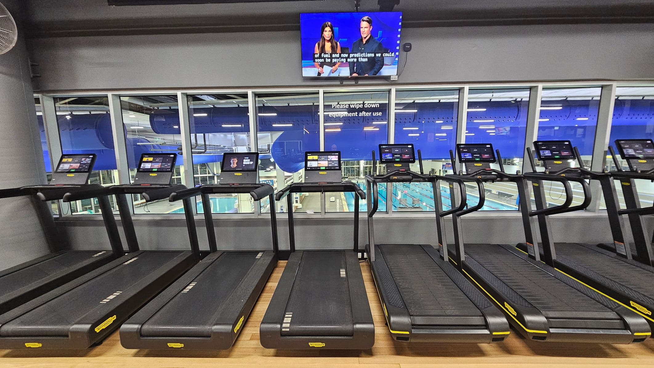 A row of treadmills with digital screens is positioned in the foreground on a wooden floor. Behind them, large windows reveal a view of an indoor swimming pool area with blue accents. Above the windows, a television screen displays a news broadcast featuring two presenters. A sign on the window reads, "Please wipe down equipment after use." The ceiling is industrial with visible ducts and lighting.