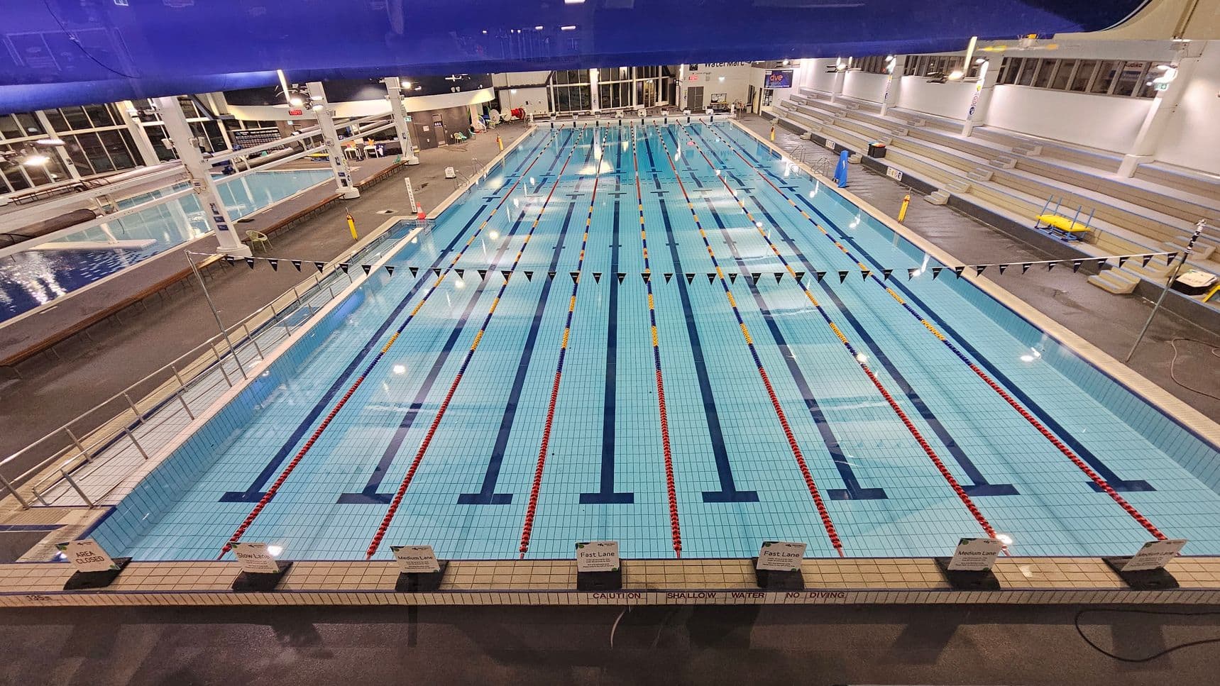 An indoor swimming pool with multiple lanes, each marked by red and yellow lane dividers. Overhead, a row of triangular flags stretches across the pool. On the right, there are tiered seating areas with benches. To the left, another smaller pool is visible. The floor surrounding the pool is dark and wet, and the ceiling is high with exposed beams and lighting fixtures. Signs at the pool's edge indicate lane speeds.