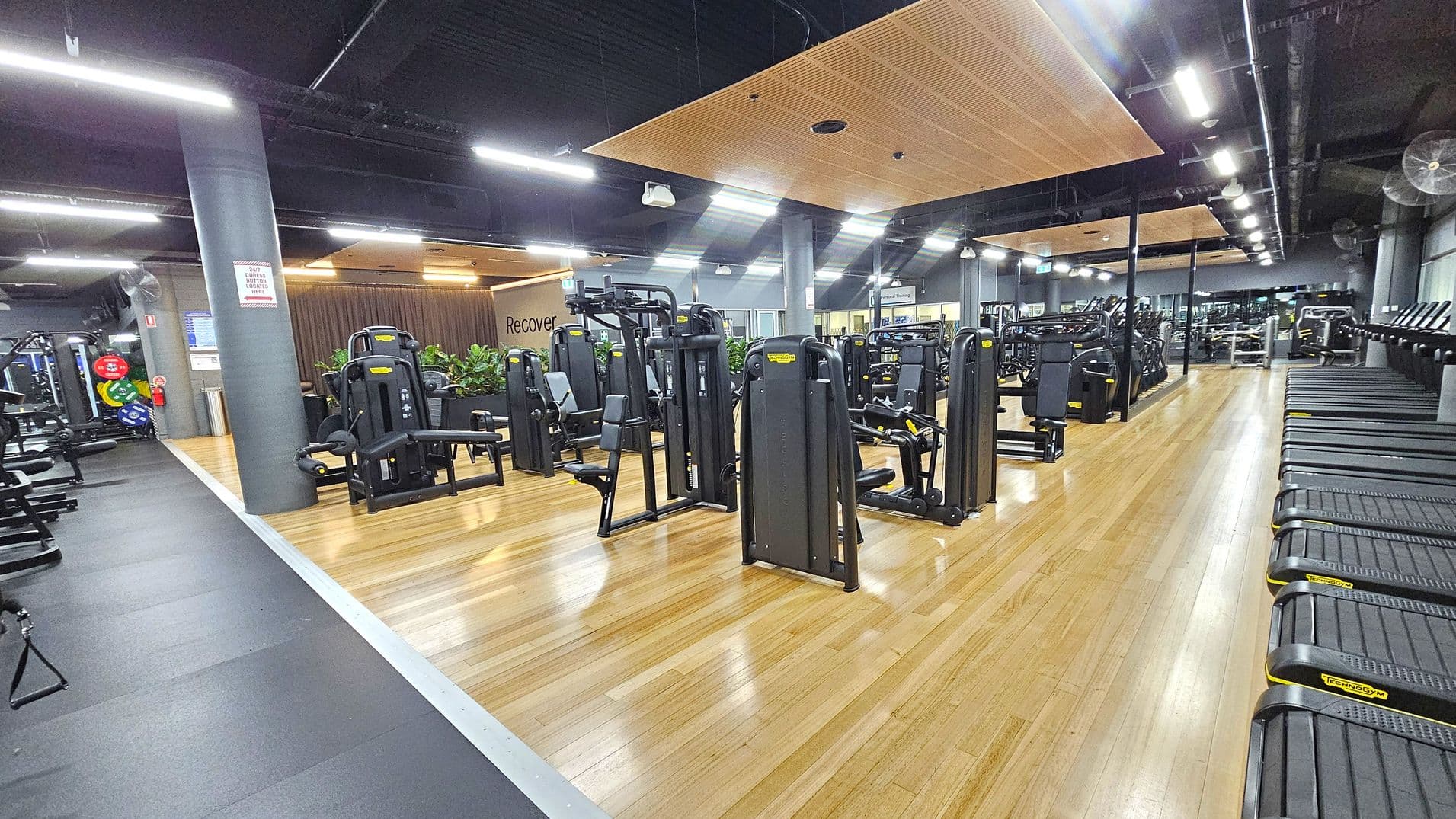 A spacious gym interior with wooden flooring. On the left, there are various weight machines and a column with a safety sign. In the centre, more exercise machines are arranged neatly, with a "Recovery" sign on the wall behind them. On the right, a row of treadmills lines the wall. The ceiling is dark with exposed beams and bright overhead lights.