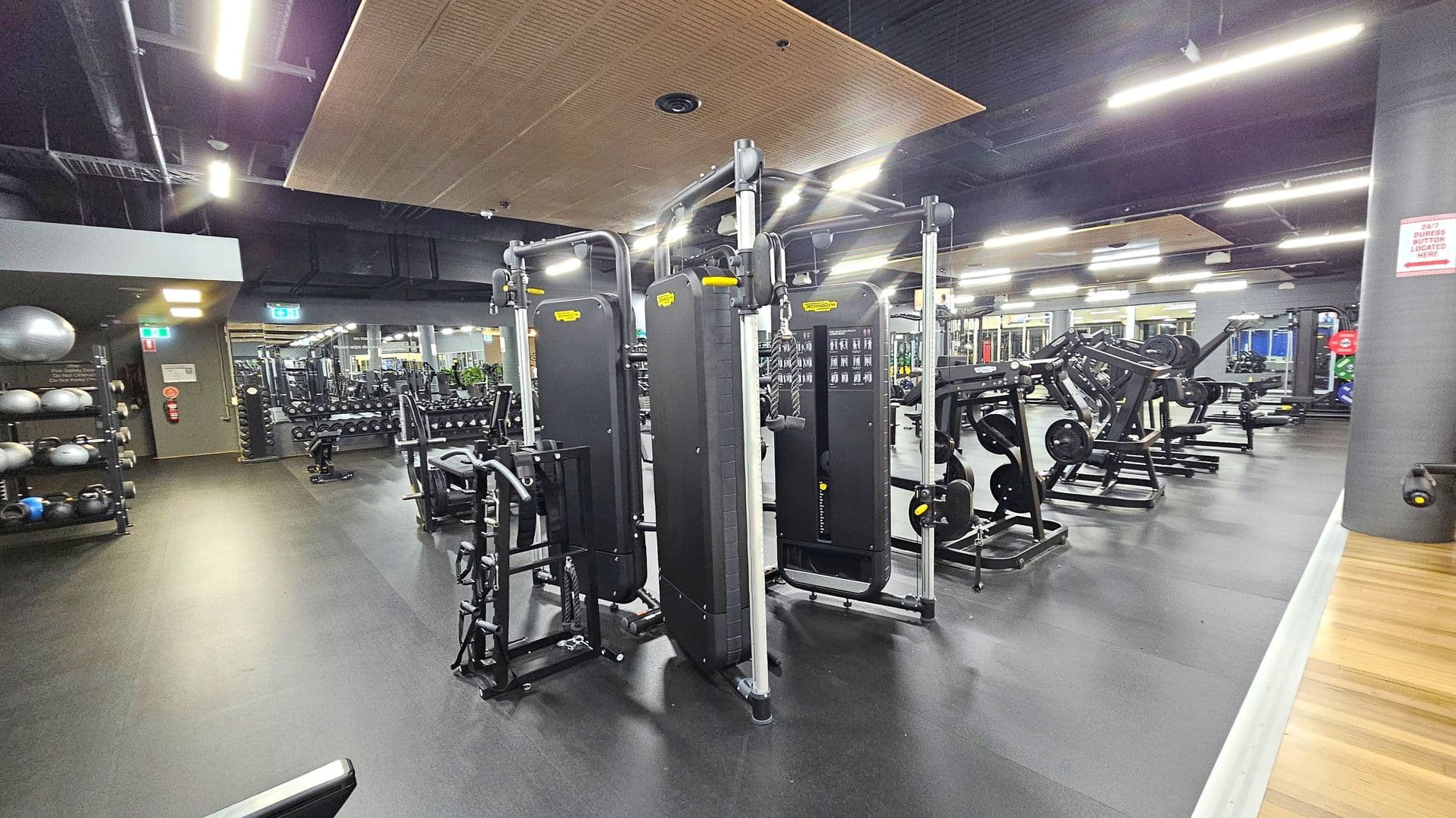 A spacious gym interior with a variety of exercise equipment. On the left, there are shelves with medicine balls and kettlebells. In the centre, several cable machines are arranged in a row. To the right, there are weight machines and benches. The floor is dark and rubberised, while the ceiling features exposed ducts and wooden panels. Bright overhead lights illuminate the space, and mirrors line the walls, reflecting the equipment.