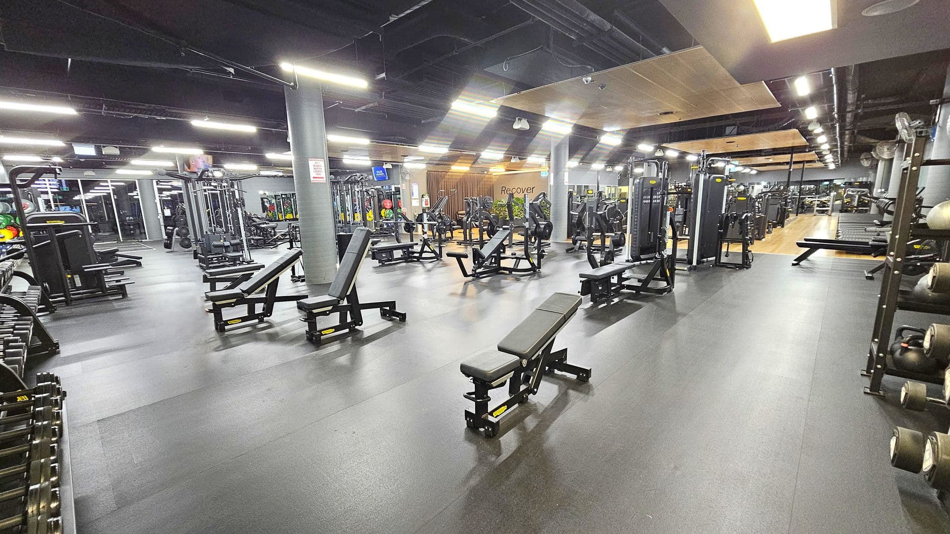 A spacious gym with various workout equipment. On the left, there are weight racks and benches. In the centre, multiple benches and weight machines are arranged neatly. On the right, more machines and a rack with medicine balls are visible. The floor is dark and smooth, while the ceiling has exposed ducts and bright lighting. In the background, a wooden panelled area with the word "Recover" is visible, along with more equipment and mirrors.