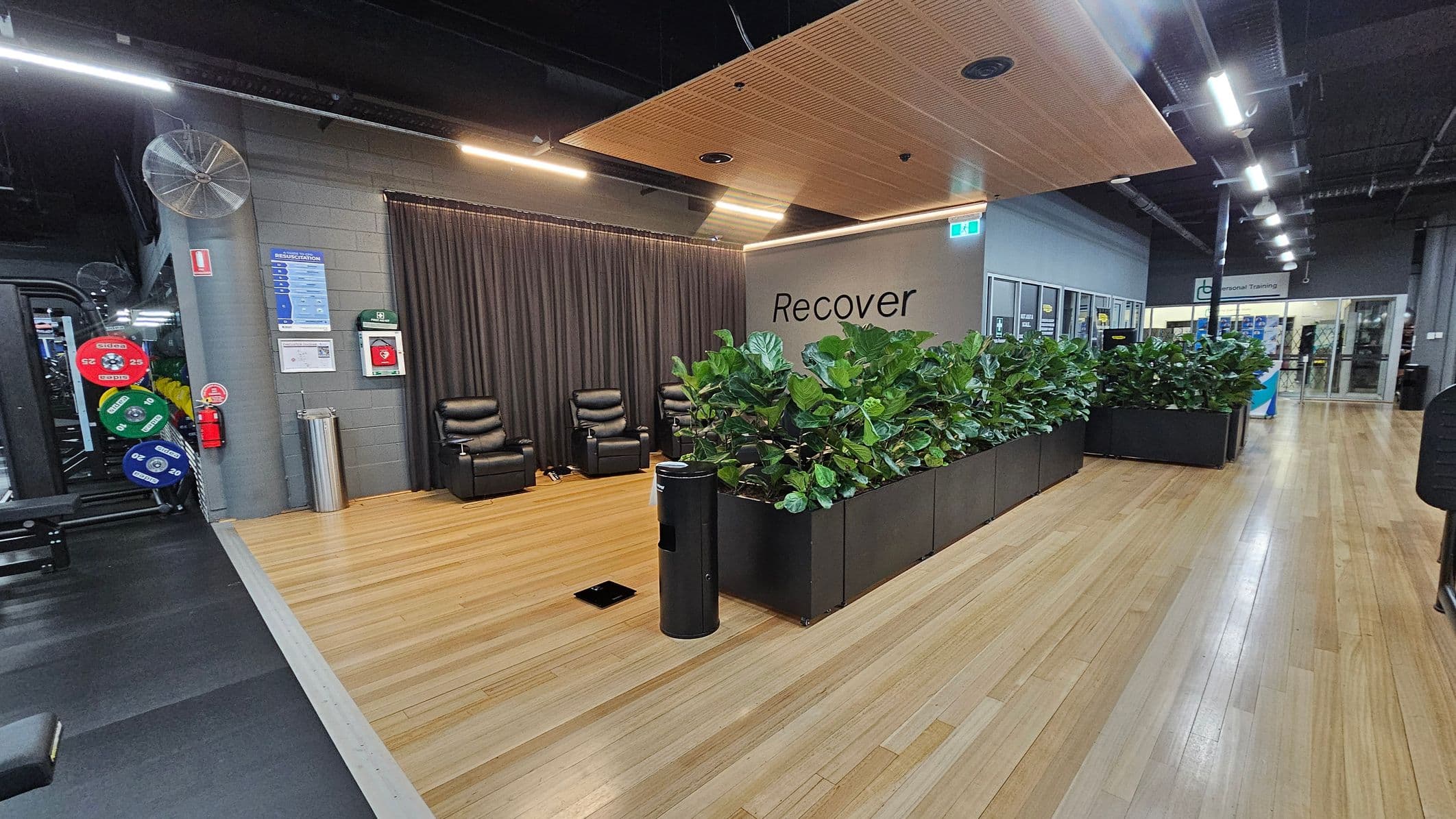 A gym recovery area with wooden flooring. On the left, there are weight plates and gym equipment. In the centre, two black recliner chairs are positioned against a dark curtain. A wall with the word "Recover" is visible, with a row of green plants in black planters in front. On the right, a corridor leads to a glass entrance with signs for personal training. The ceiling has wooden panels and exposed lighting fixtures.