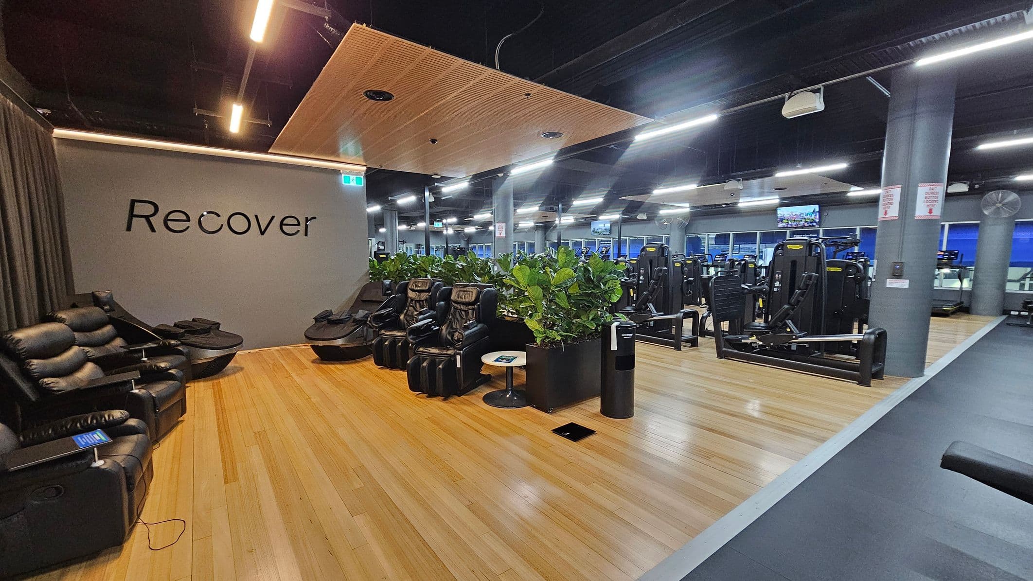 A gym interior with a recovery area on the left featuring black massage chairs and a grey wall with the word "Recover". The floor is wooden, and a small round table is nearby. In the centre, there are potted plants separating the recovery area from the gym equipment. On the right, various exercise machines are visible. The ceiling is dark with exposed fixtures and bright linear lights.