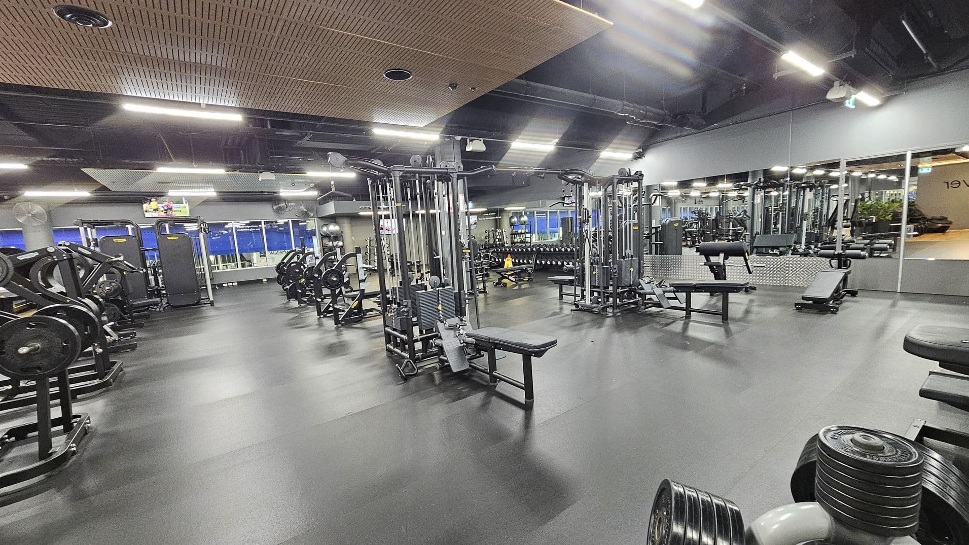 A spacious gym with a variety of exercise equipment. On the left, there are weight machines and benches. In the centre, there are cable machines and benches. The right side features a mirrored wall reflecting the gym equipment and a row of dumbbells. The floor is dark and rubberised, while the ceiling has a mix of exposed ducts and wooden panels. Bright overhead lights illuminate the space, and a TV is mounted on the wall in the background.