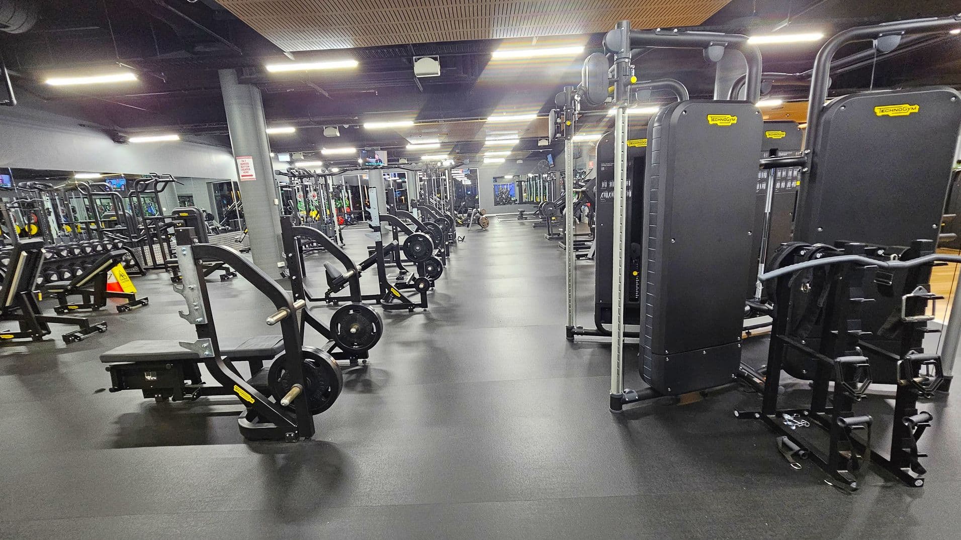 A spacious gym with various exercise machines. On the left, there are weight benches and racks with dumbbells. In the centre, multiple weightlifting machines are aligned in rows. On the right, there are large, upright fitness machines. The floor is covered with dark rubber mats, and the ceiling has exposed pipes and bright fluorescent lights. A grey pillar stands near the centre, and a caution sign is visible on the left.