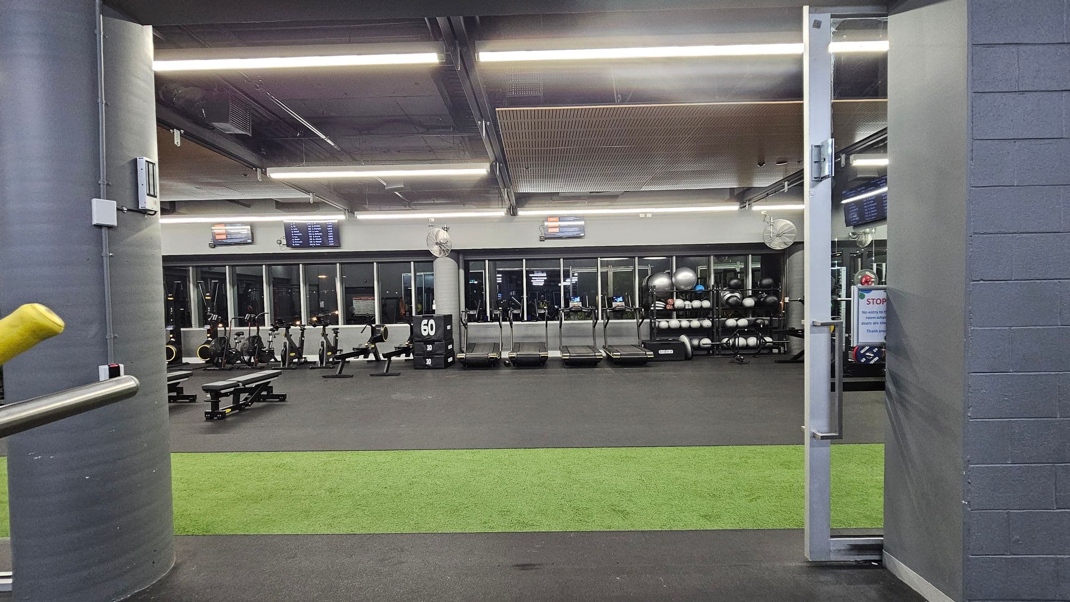 A gym interior with a grey and green colour scheme. In the foreground, a green artificial turf strip is visible on the floor. The background features a row of treadmills and exercise bikes against a wall with large windows. To the right, shelves hold exercise balls and other equipment. The ceiling has exposed beams and fluorescent lighting. A grey pillar is on the left, and a partially open door is on the right.
