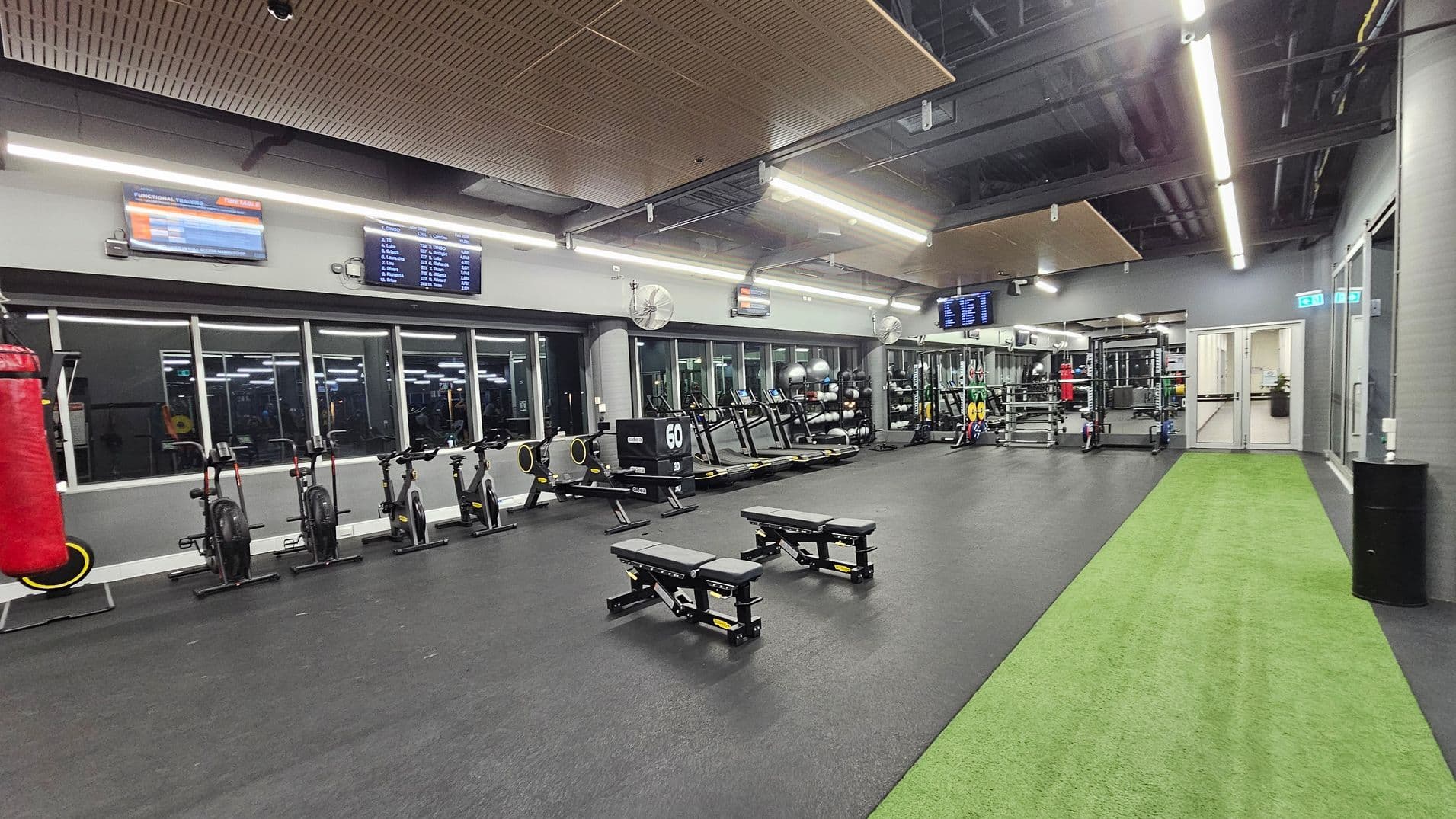 A spacious gym interior with a black rubber floor and a green artificial turf strip on the right. On the left, there are stationary bikes and a red punching bag. In the centre, two adjustable benches are positioned. The back wall features large mirrors and windows, with various gym equipment like treadmills and weights. The ceiling has exposed ducts and wooden panels, with bright fluorescent lighting. Several screens displaying information are mounted on the walls.