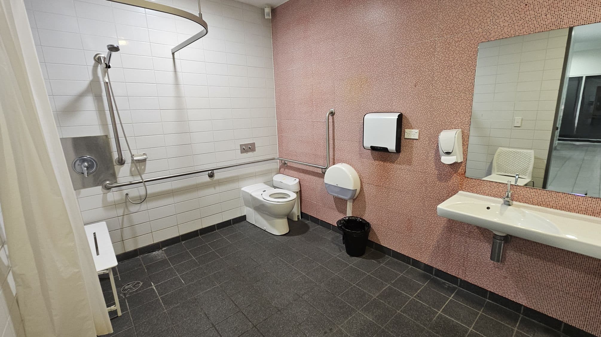 A spacious accessible bathroom with a dark tiled floor. On the left, there is a shower area with a handheld showerhead, grab bars, and a white shower seat. The back wall has white tiles. In the centre, a toilet with grab bars on both sides is visible. On the right, a pink-tiled wall features a large mirror above a white sink. A paper towel dispenser and soap dispenser are mounted on the wall. A black waste bin is on the floor. The ceiling is plain white.