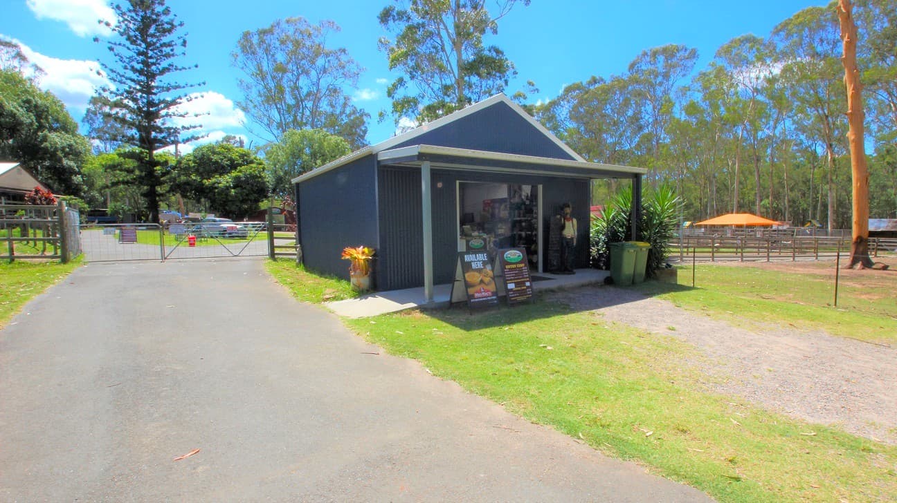 Outside the ticket office are two metal banners. One banner has images of snacks on it while the other banner shows the entrance charges for the White Ridge Farm. Next to the ticket office is a large green bush. At the door to the ticket office is a male manikin dressed like a cowboy in a polka-dotted white shirt, black trousers, a cowboy hat and a red scarf on the neck.