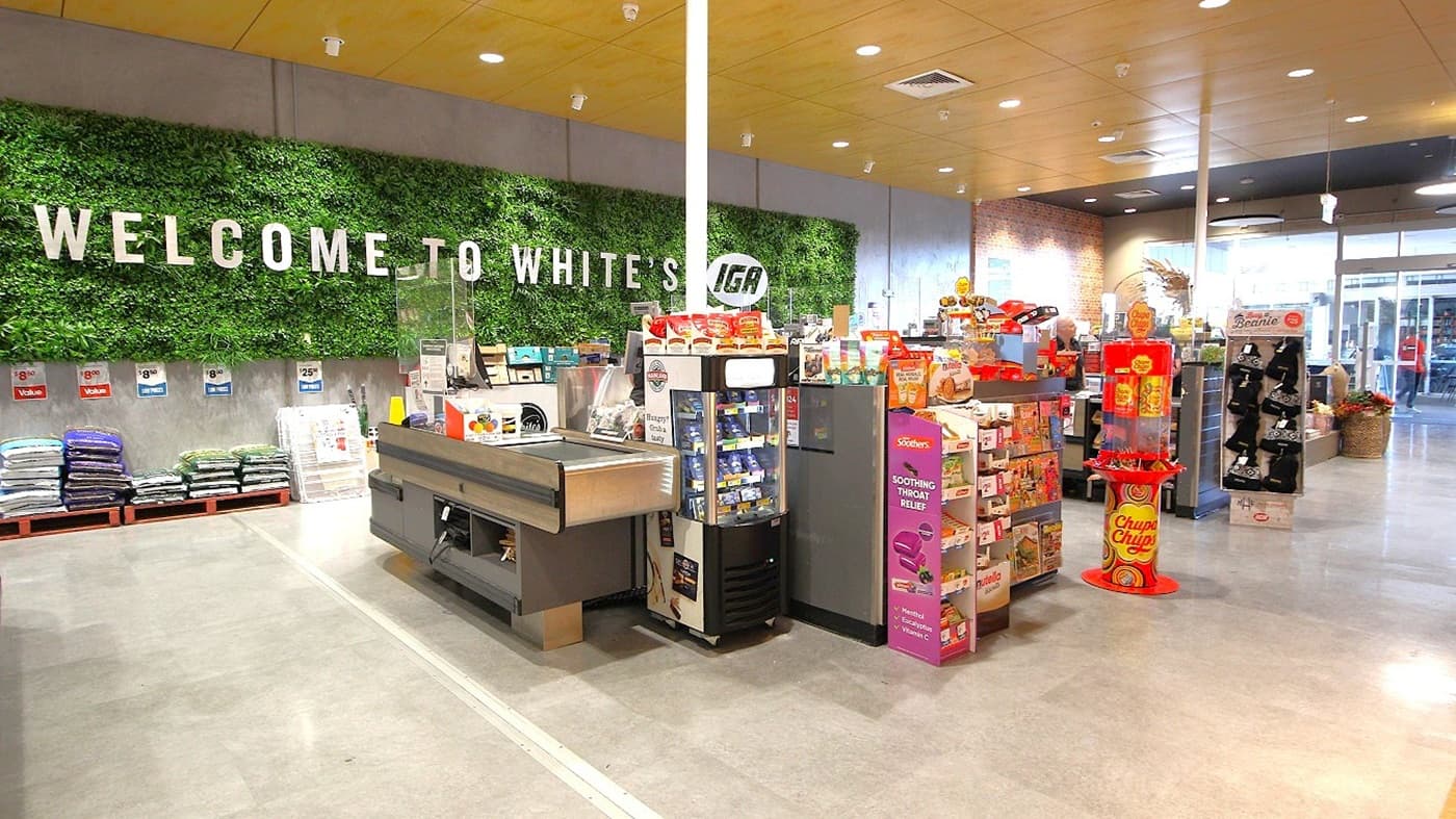 In the foreground, there are checkout counters with a variety of products, including snacks and beverages on display. To the left, neatly stacked bags of mulch and soil are arranged on wooden pallets. On the right, several stands showcase items like sweets and health products. Behind the counters, a green wall features large letters saying "WELCOME TO WHITE'S IGA" among artificial plants. The flooring is a smooth, grey concrete, and the ceiling is a flat, light wood with recessed lights.