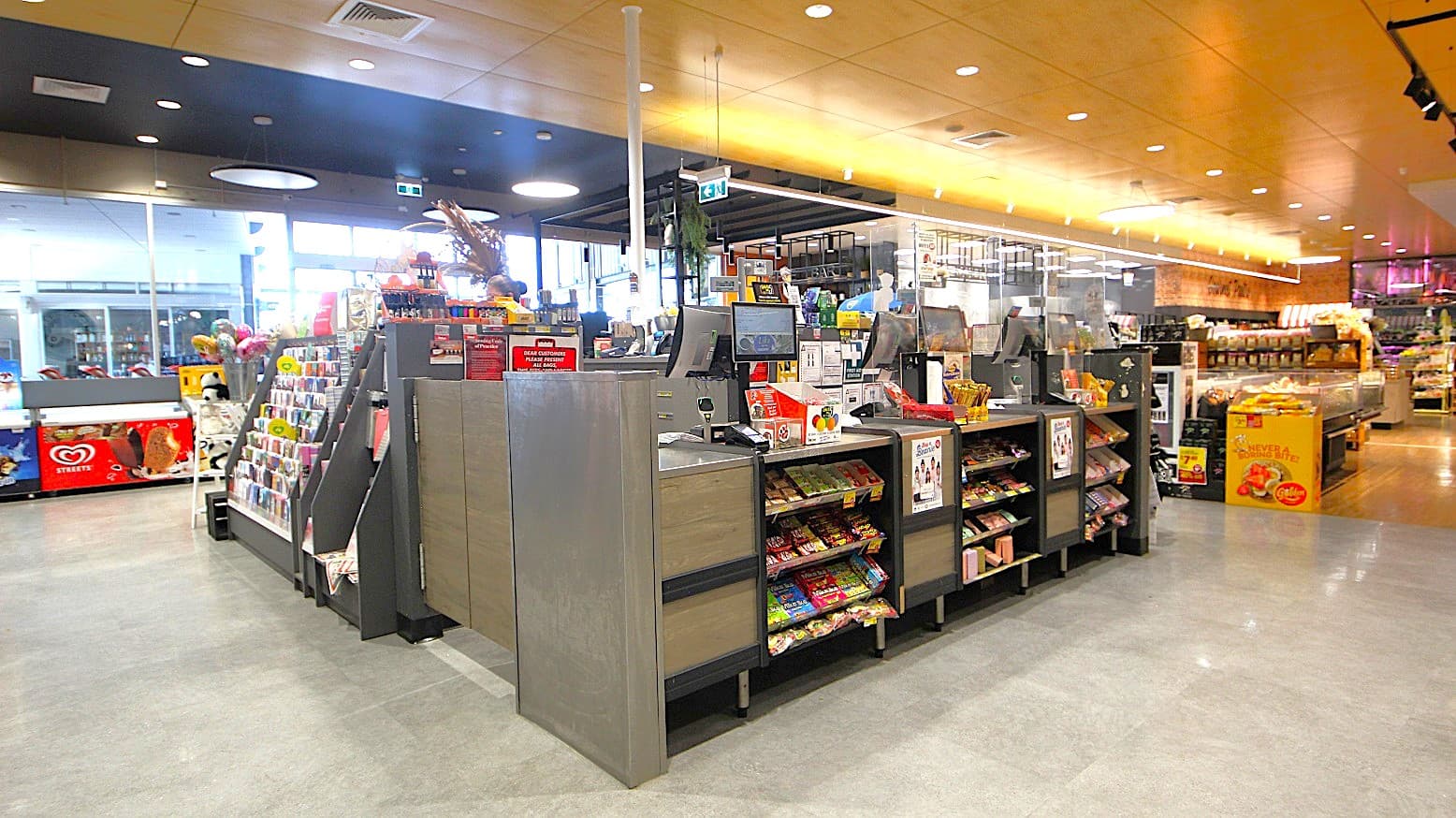 In the foreground, multiple checkout counters are arranged, each equipped with a computer and surrounded by displays of snacks and candies. To the left, an upright display stand showcases greeting cards and small gifts. In the background, large windows allow natural light into the space, and shelves filled with various grocery items and ready-made meals are visible. The flooring is smooth grey concrete, while the ceiling features wooden panels with circular light fixtures.