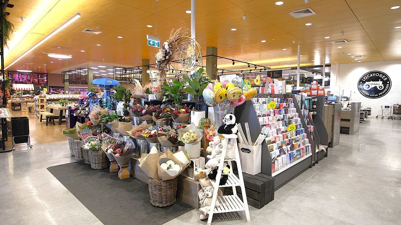 In the foreground, a vibrant flower display takes centre stage, featuring various bouquets wrapped in brown paper, stainless steel buckets filled with plants, and plush toys arranged decoratively. To the right, racks showcase greeting cards and small gifts. In the background, checkout counters are visible, alongside additional shelving filled with produce and groceries. The flooring is grey concrete, while the ceiling features light wooden panels with recessed lighting and suspended fixtures.