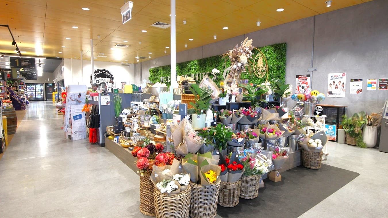 In the foreground, a large floral display features bouquets in various colours, arranged in brown paper and placed in wicker baskets. To the right, there is a range of household items and gifts on display, alongside additional plants. Behind the floral section, a green wall displays the words "WELCOME TO WHITE'S IGA" in bold lettering. To the left, checkout counters are visible, and various grocery items are stocked on shelves.
