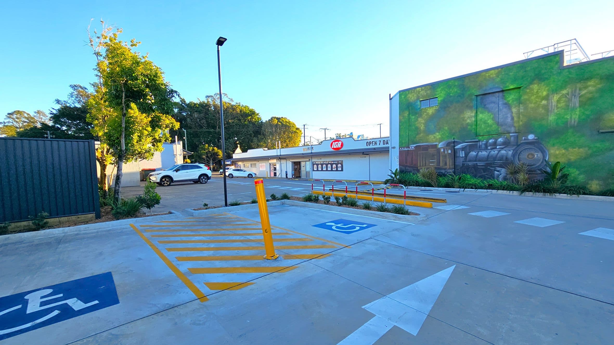 A car park with a designated disabled parking space marked in blue on the ground. A yellow bollard and striped lines are nearby. On the right, a large mural of a steam train is painted on a building wall. In the background, a white car is parked near a small shopping area with an IGA store. Trees and a lamppost are visible on the left, and the ground is paved with concrete.