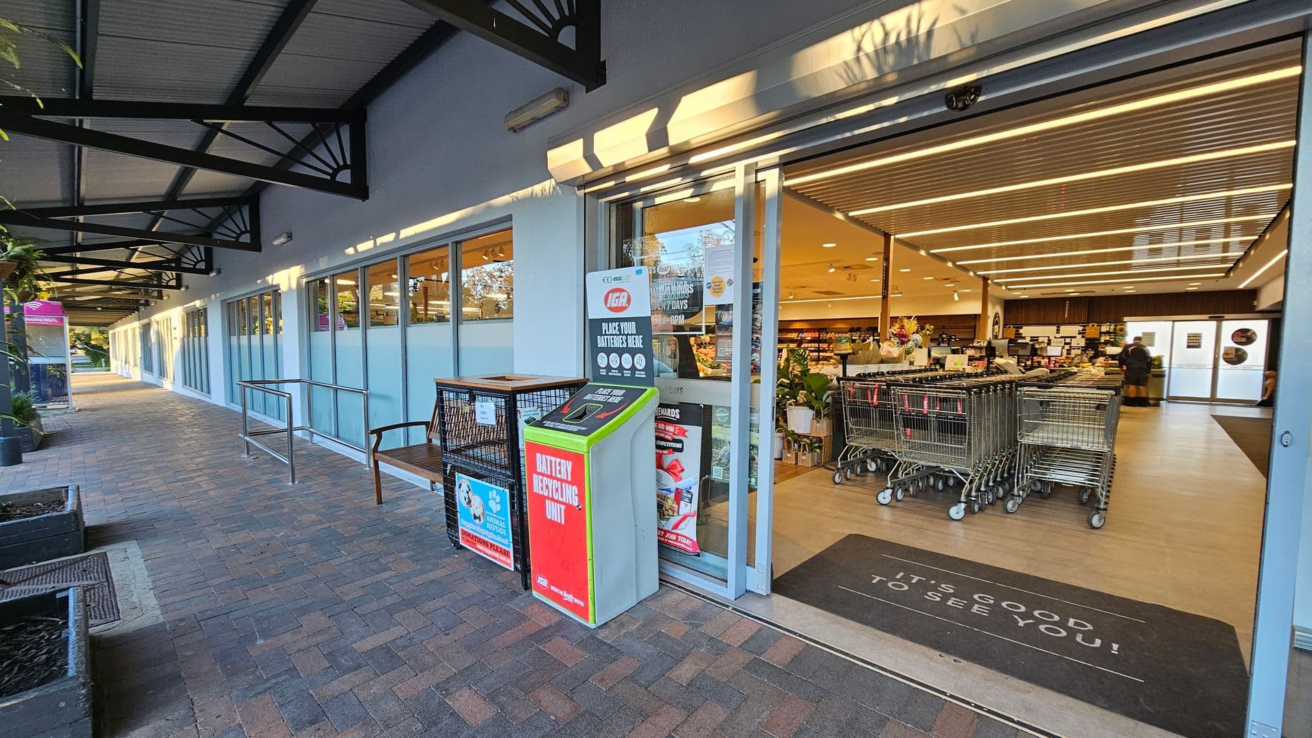 A supermarket entrance with a row of shopping trolleys on the right. The floor inside is light-coloured, contrasting with the brick pavement outside. A mat at the entrance reads "IT'S GOOD TO SEE YOU!" On the left, a battery recycling unit and a donation bin are placed near the glass doors. The ceiling inside features parallel strip lighting. The exterior has a covered walkway with metal supports and large windows.