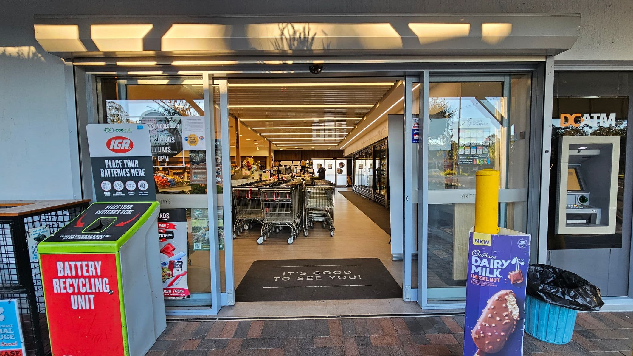 Automatic sliding glass doors lead into a supermarket. On the left, a battery recycling unit with a sign above it. Inside, shopping trolleys are lined up. On the right, an ATM machine is visible next to a bin with a promotional sign for a chocolate ice cream. The floor is tiled, and a welcome mat reads "IT'S GOOD TO SEE YOU!" The ceiling has a series of parallel lights.