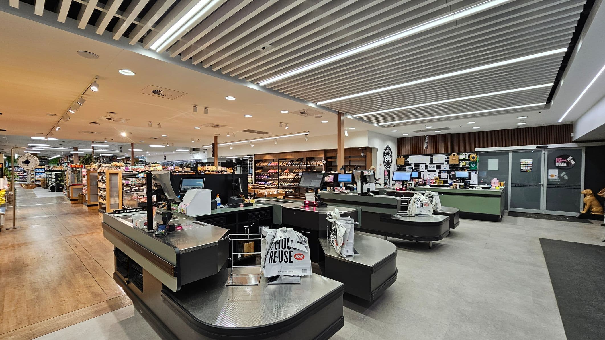 Checkout area of a supermarket with two checkout counters in the foreground, each equipped with a monitor and a card reader. Reusable shopping bags are placed on the counters. In the background, shelves are stocked with various products, including fruits and bakery items. The ceiling features a grid of linear lights, and the floor is a mix of light and dark tiles. The store is well-lit, creating a bright and organised atmosphere.