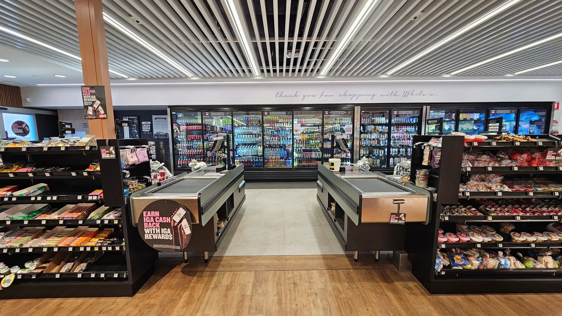A supermarket checkout area with two conveyor belts in the centre. Behind them, a wall of refrigerated drink displays is visible. On the left and right, shelves are stocked with various snacks and packaged goods. The floor is wooden, and the ceiling features a grid of linear lights. A sign above the drink displays reads, "thank you for shopping with White's."