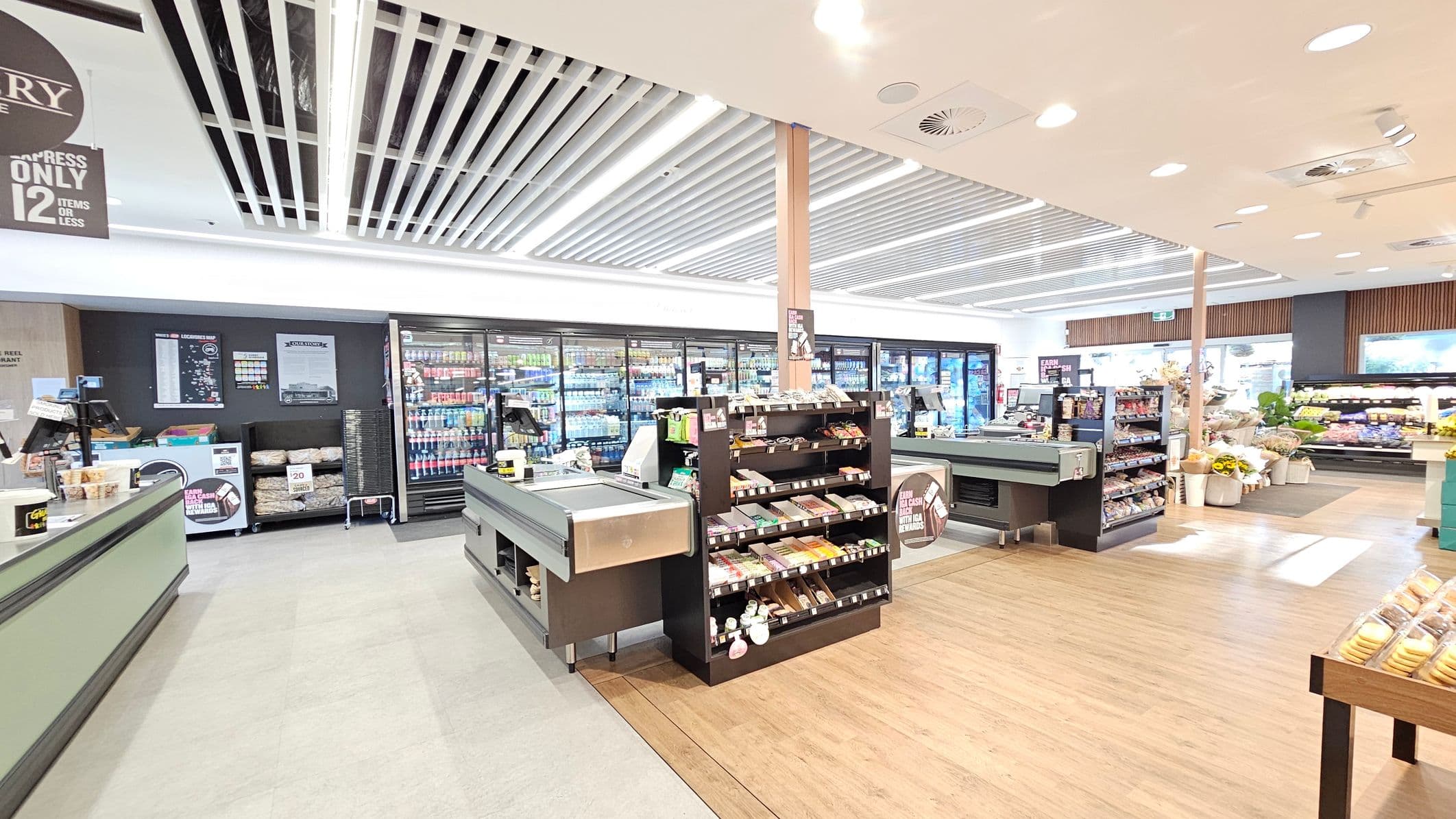 A supermarket interior with checkout counters in the foreground, featuring shelves stocked with snacks and small items. The background displays refrigerated sections filled with beverages. To the right, there are displays of fresh produce and flowers. The ceiling has a grid pattern with recessed lighting, and the floor is a mix of light wood and grey tiles. A sign above indicates an express checkout lane for 12 items or less.