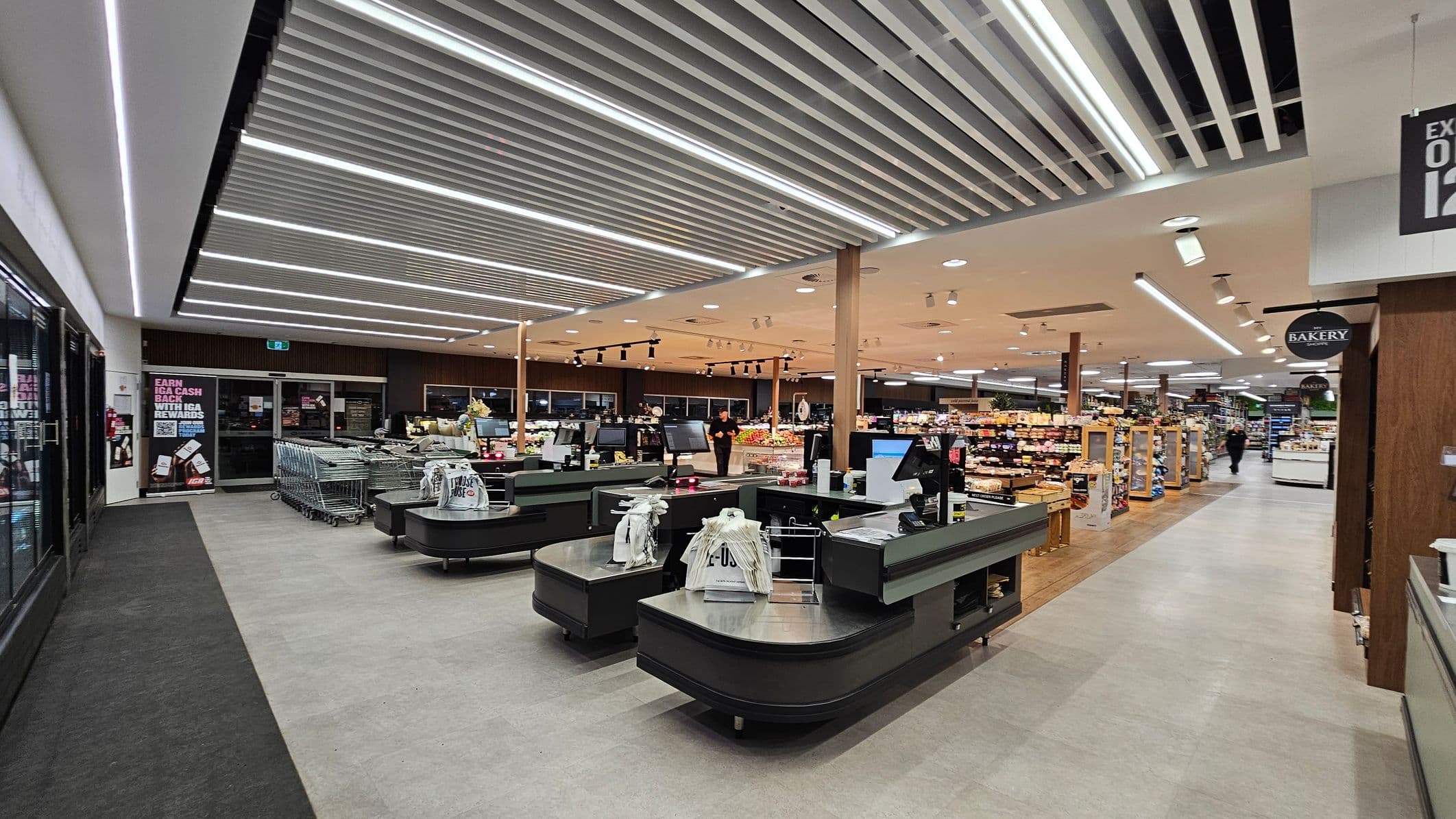 A supermarket interior with checkout counters in the foreground, featuring black surfaces and bagged items. On the left, there are shopping trolleys and a glass door with promotional posters. The right side shows aisles filled with various products, and a "Bakery" sign is visible. The ceiling has a modern design with linear lights, and the floor is a mix of grey and wood textures. The background includes more aisles and a few shoppers.