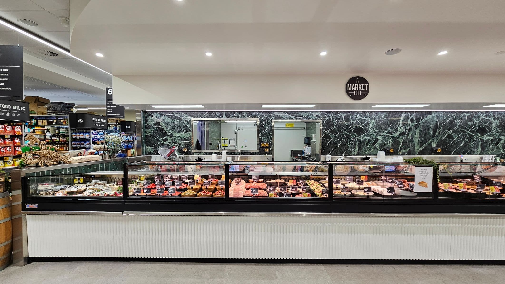 A well-lit supermarket interior with wooden flooring. In the centre, there's a refrigerated display case filled with packaged meats. To the left, a small dining area with a table and chairs is visible, along with shelves of products and decorative plants. On the right, a long refrigerated section displays fresh produce. The ceiling features large circular lights, and the background shows more shelves stocked with various goods.