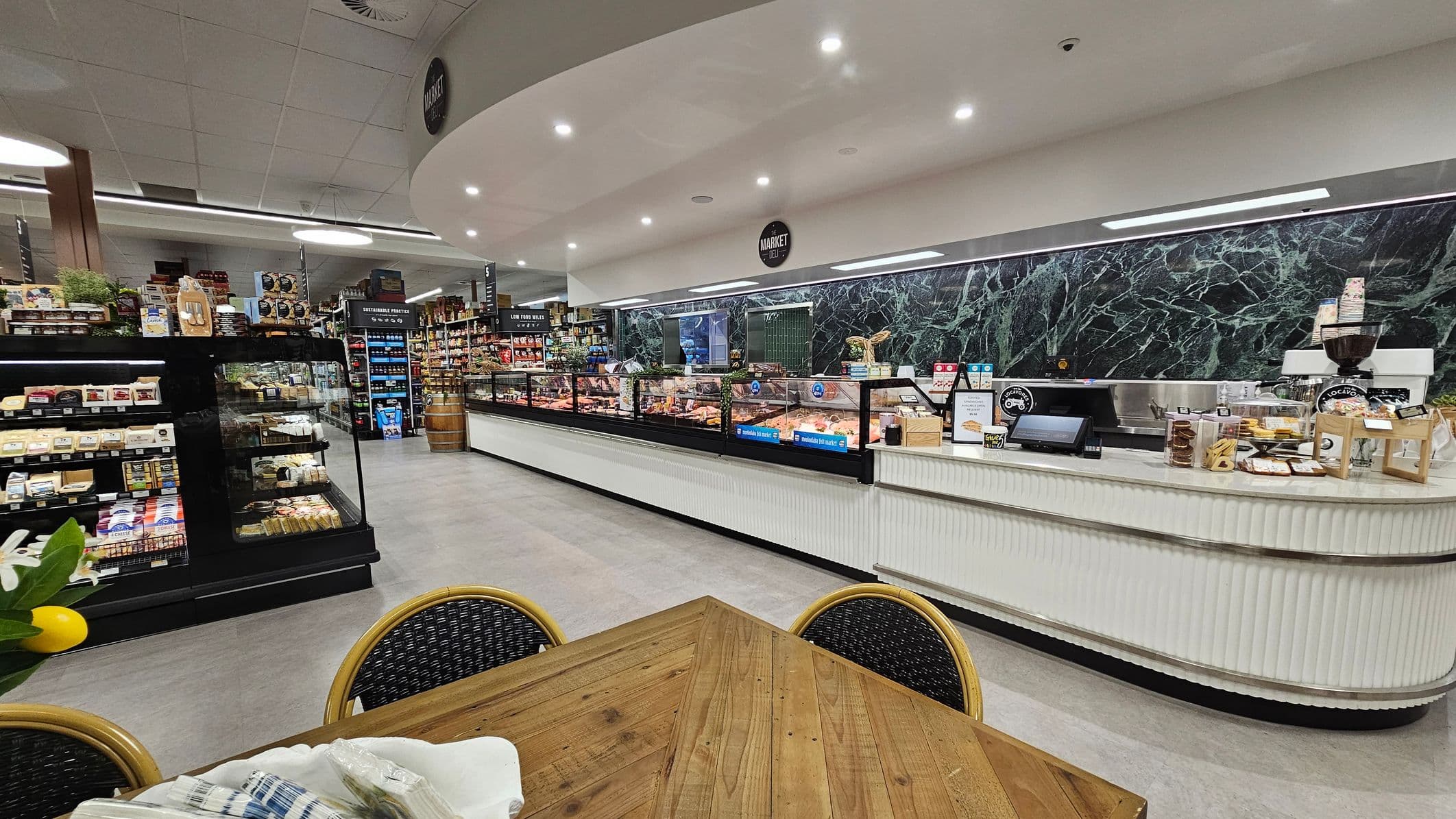 A deli section in a supermarket with a long, white counter on the right, featuring a green marble backdrop. Various products and a coffee machine are displayed on the counter. In the middle, a glass display case contains meats and cheeses. On the left, shelves are stocked with packaged goods. In the foreground, a wooden table with wicker chairs is visible. The ceiling is fitted with recessed lighting and a grid pattern.