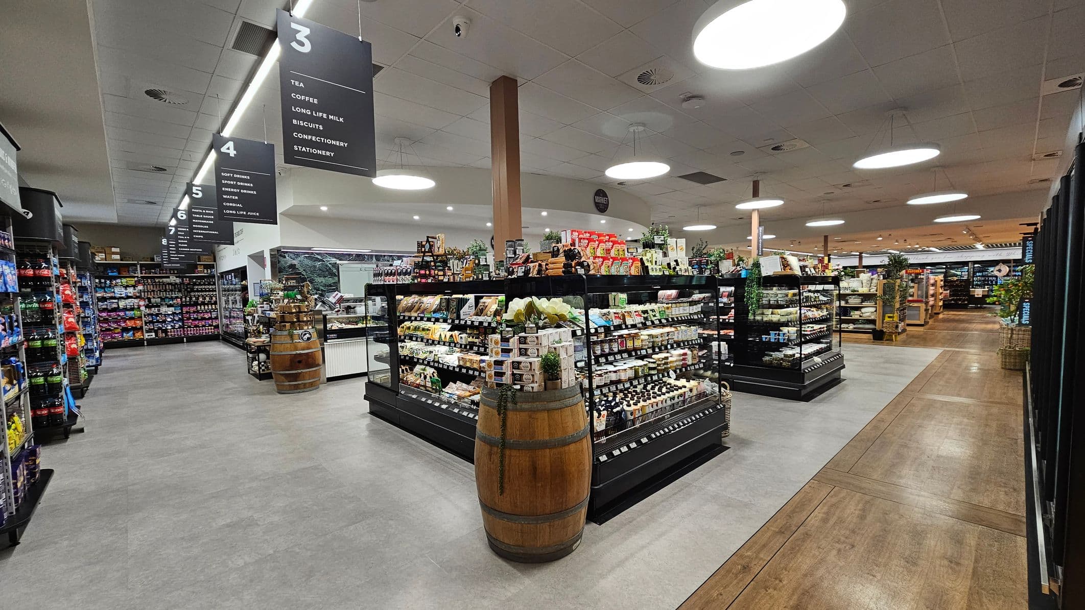 A supermarket interior with wide aisles and overhead signs indicating sections like tea, coffee, and confectionery. Shelves are stocked with various products, including drinks and snacks. In the centre, a wooden barrel is used for display. The floor transitions from grey tiles to wood, and the ceiling features large circular lights. The space is well-organised, with clear signage and neatly arranged products.