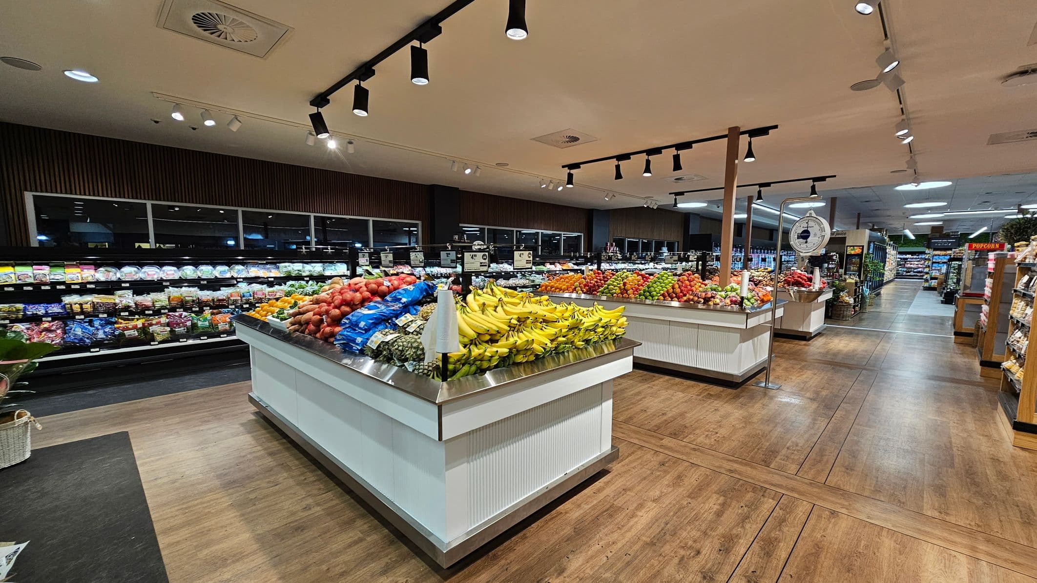A supermarket interior with wooden flooring and a light-coloured ceiling featuring spotlights. In the foreground, there are displays of fresh produce, including bananas, apples, and other fruits. To the left, refrigerated shelves hold packaged goods. In the background, more produce displays and a scale are visible. A popcorn sign is on the right, near shelves with various products.