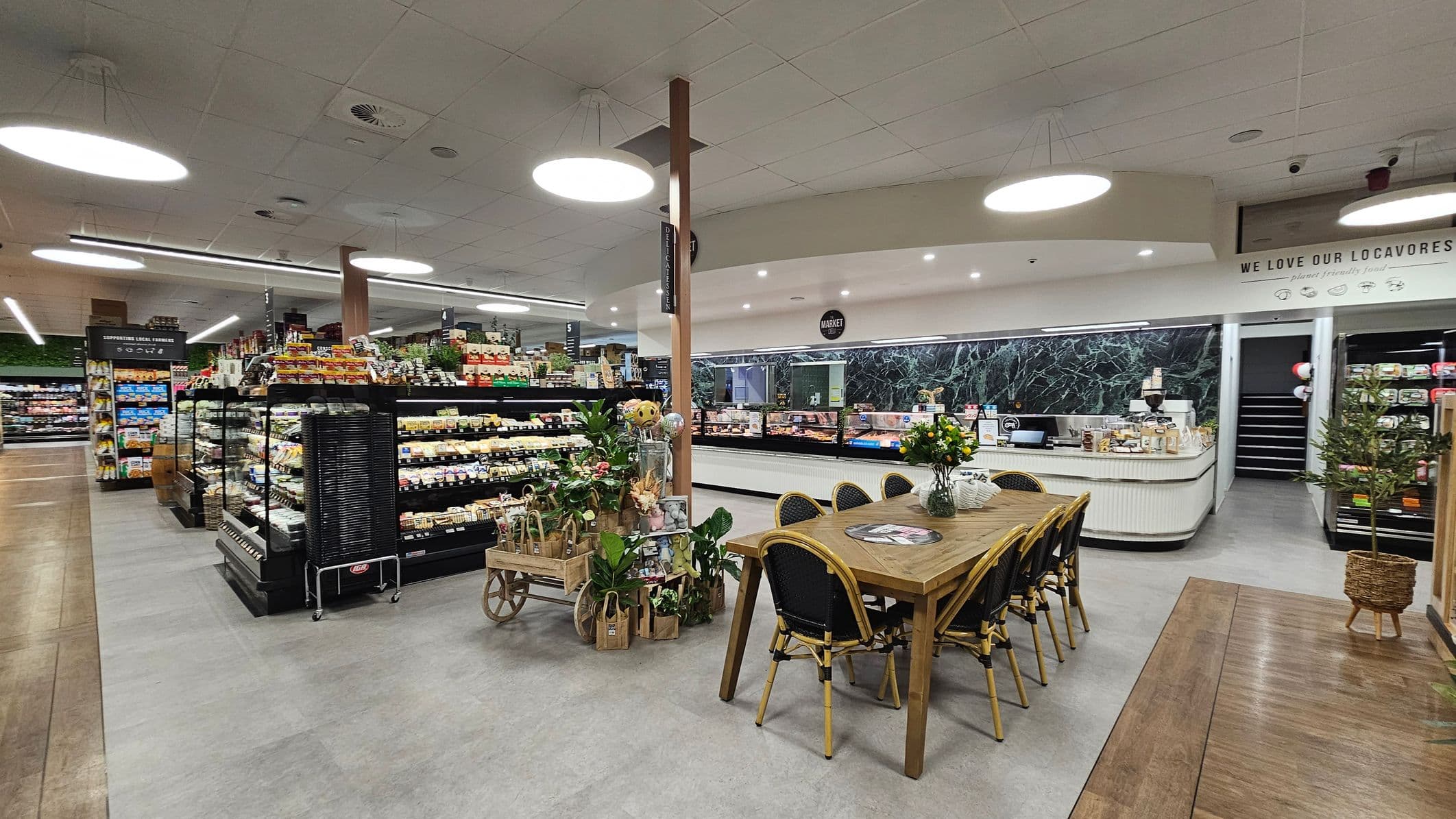 A supermarket interior with a modern design. On the left, shelves are stocked with various packaged goods. In the centre, a wooden table with chairs is adorned with a vase of flowers. Nearby, a decorative cart holds plants and gift bags. The right side features a counter with a coffee machine and a display case, backed by a dark green marble wall. The ceiling is fitted with large circular lights, and the floor is a mix of wood and grey tiles.