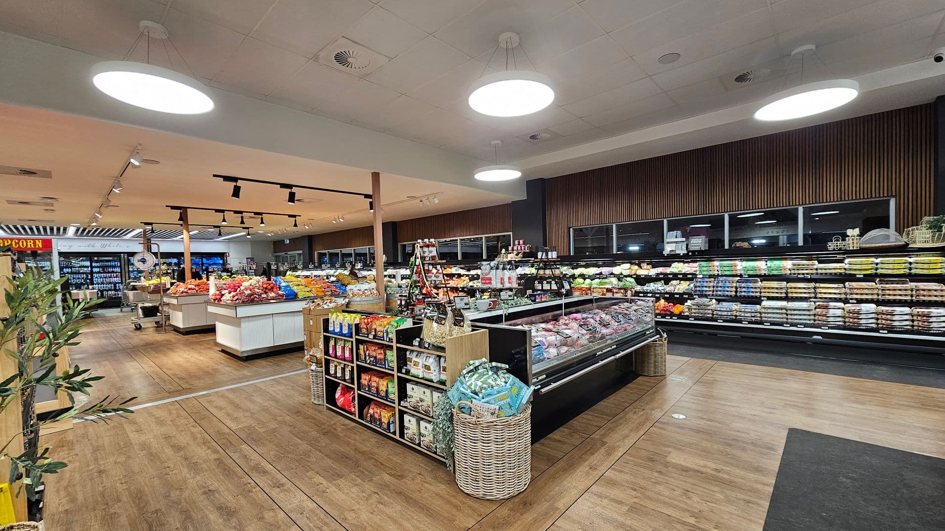 A supermarket interior with wooden flooring and a ceiling featuring circular lights. On the left, there are shelves with various snacks and a popcorn sign. In the centre, displays of fresh produce like fruits and vegetables are visible. On the right, refrigerated sections contain dairy products and eggs. A wicker basket is placed near the shelves, and the area is well-lit with a modern design.