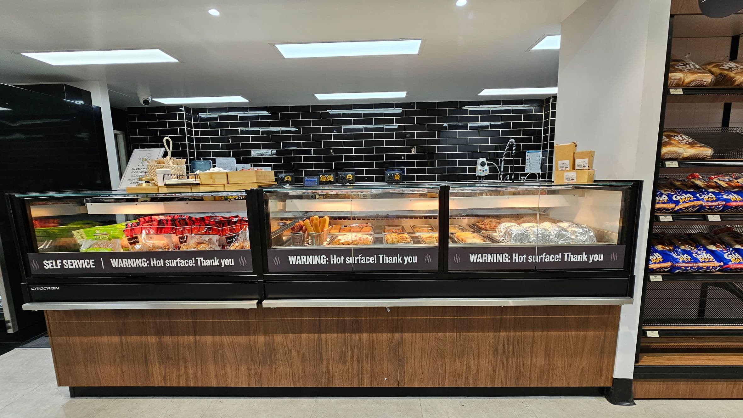 A self-service food counter with a wooden base and glass display, featuring a warning about hot surfaces. Inside, various food items are visible, including packaged goods and baked items. Behind the counter, a black tiled wall with white grout and a sink area with utensils and boxes. On the right, shelves stocked with bread and packaged snacks. The ceiling has recessed lighting, and the floor is light-coloured.