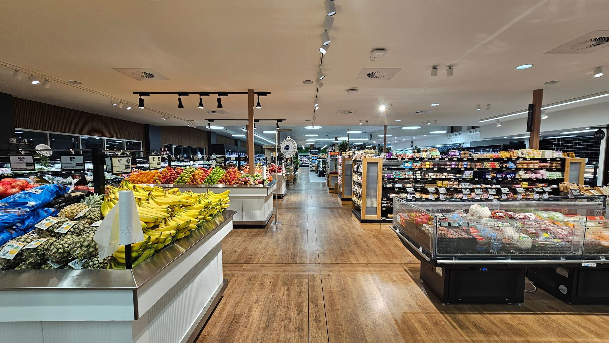 A supermarket interior with a wooden floor and a ceiling featuring recessed lighting. On the left, there is a display of fresh produce including bananas, pineapples, and various fruits. In the centre, a scale is visible, and the aisle extends into the distance. On the right, there is a refrigerated section with packaged goods and shelves stocked with various items. The background shows more aisles and shelves filled with products.