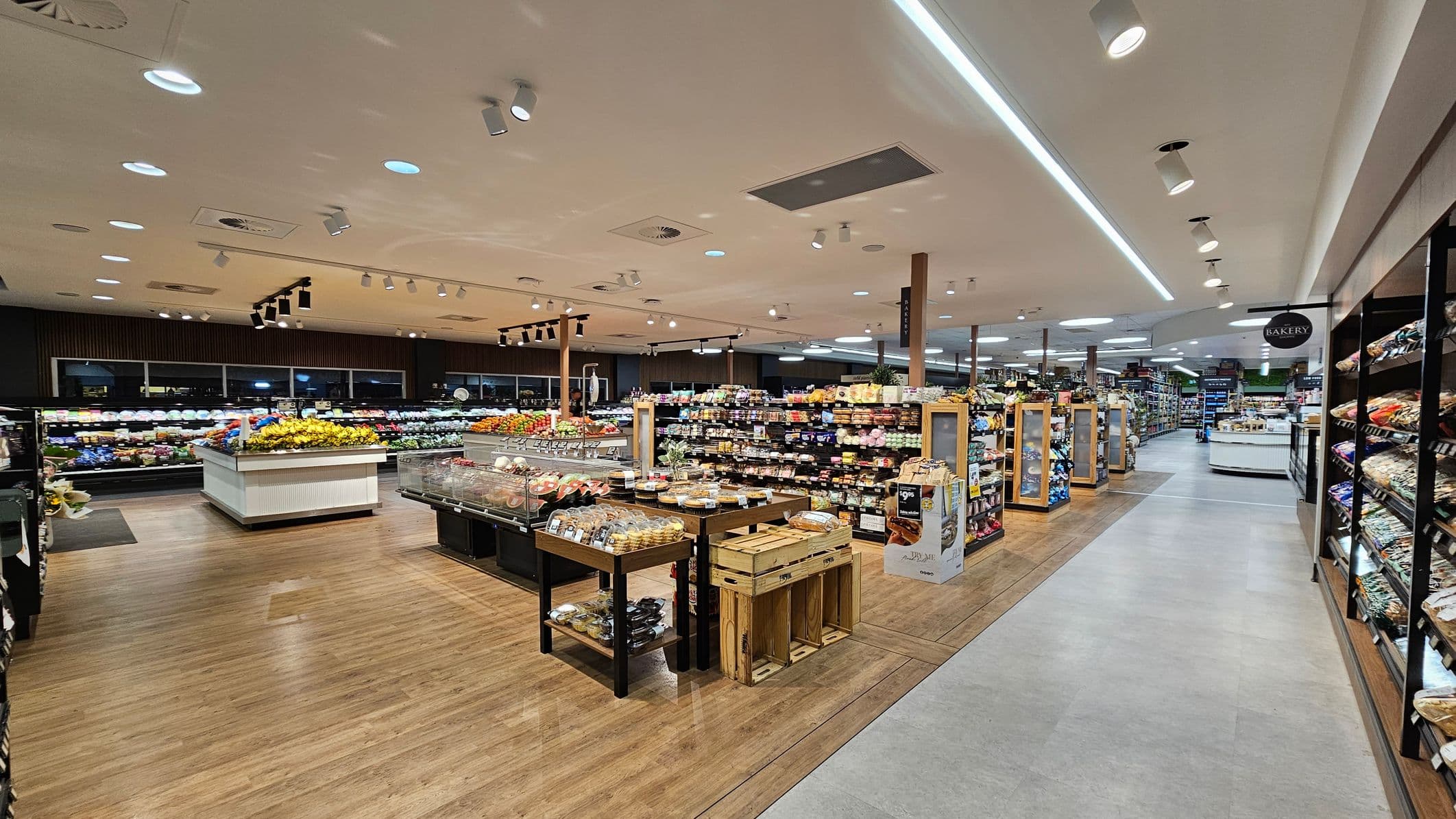 A spacious supermarket interior with a wooden floor and a light-coloured ceiling featuring recessed lighting. On the left, there are displays of fresh produce, including fruits and vegetables. In the centre, there are tables with baked goods and packaged items. The right side has shelves stocked with various products. A sign indicating the bakery section is visible. The aisles are wide, allowing for easy navigation.