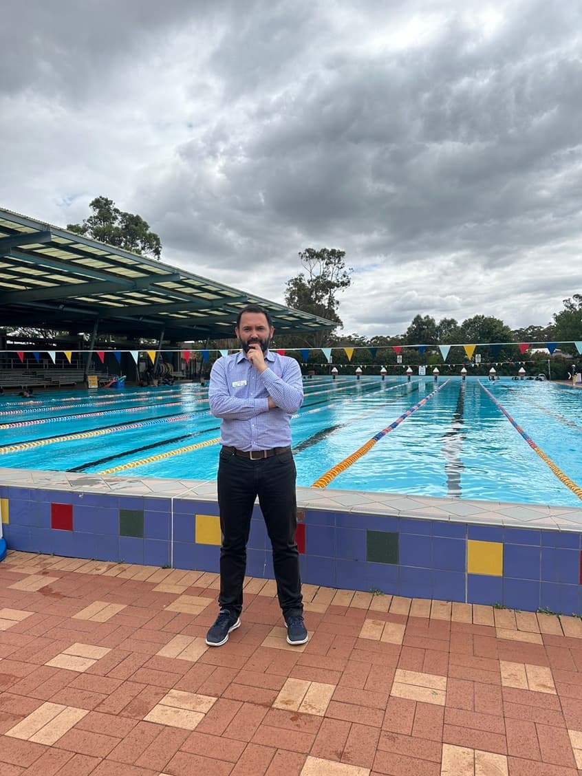 A man stands poolside with arms crossed at an outdoor swimming complex.