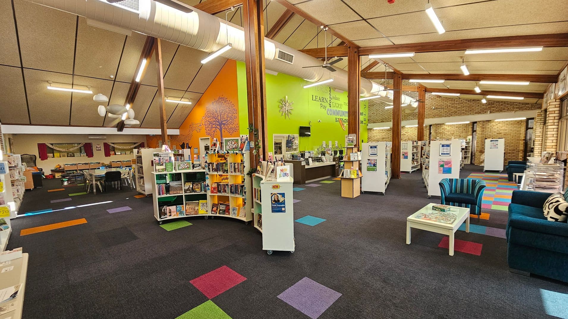 A library interior with colourful carpet tiles in shades of blue, green, red, and purple. On the left, there are bookshelves filled with books and a seating area with tables and chairs. In the centre, more bookshelves display "New Books" with a vibrant orange wall featuring a tree mural. The right side has a seating area with striped and solid blue chairs around a white table. The ceiling is high with exposed beams and ductwork, and the walls are a mix of bright green and brick.