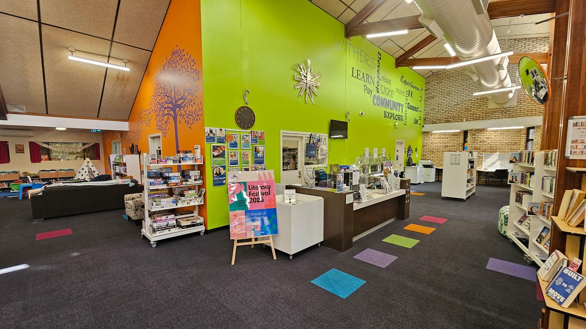 A library interior with a vibrant design. On the left, an orange wall features a purple tree mural and a clock. Below, a shelf holds board games. A black sofa and a small teepee are visible in the background. The right side has a green wall with motivational words and a reception desk with clear dividers. Bookshelves are arranged neatly, and colourful square patterns decorate the dark carpeted floor. The ceiling is high with exposed ducts and bright lighting.