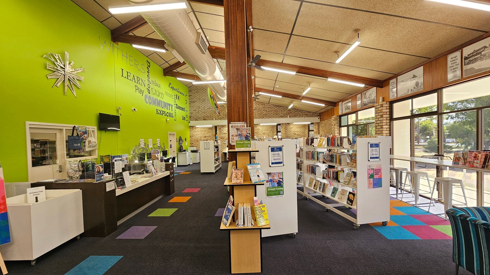A library interior with a bright green wall on the left featuring motivational words and a metallic star decoration. Below, a counter with brochures and a small TV. In the centre, a wooden pillar and a display stand with books. To the right, bookshelves on wheels and a row of high stools by large windows. The floor is carpeted with colourful square patterns, and the ceiling has exposed beams and fluorescent lights.