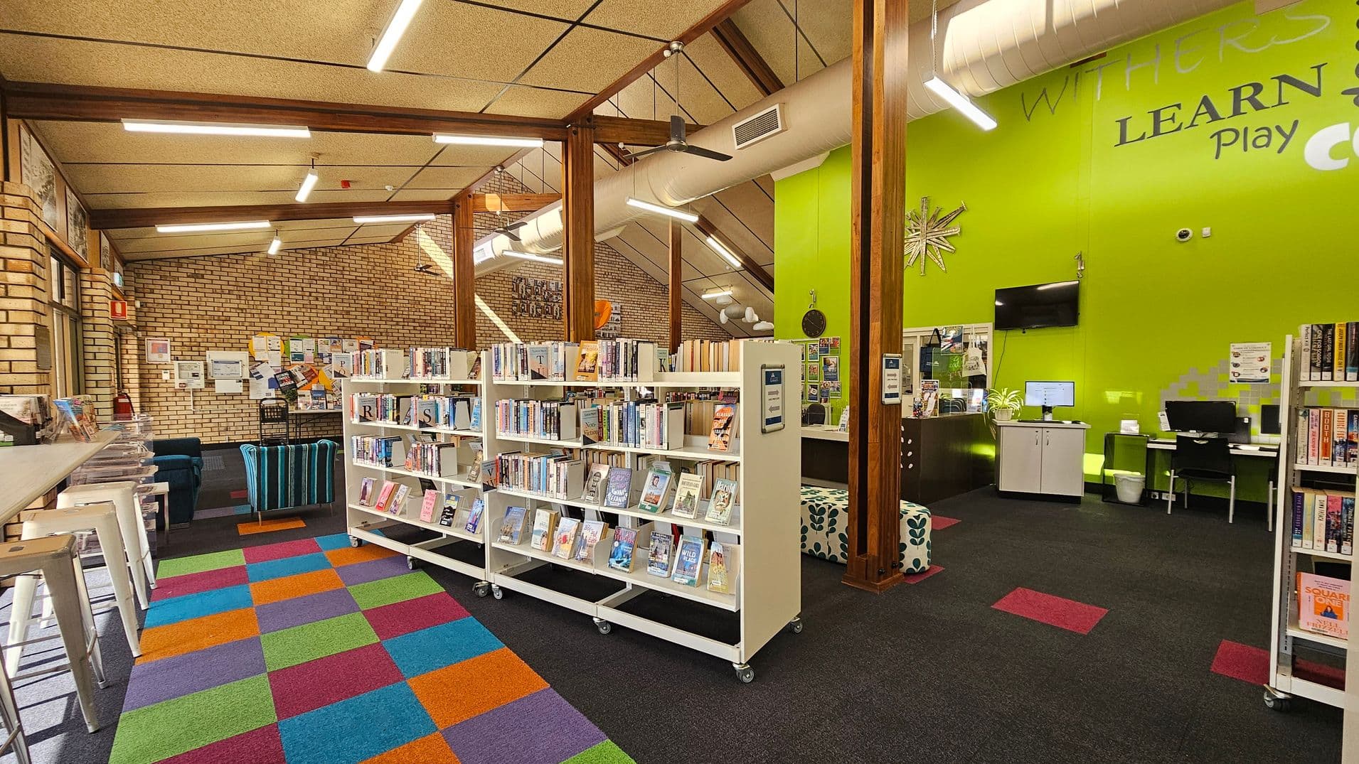 A library interior with a colourful, checkered carpet in the foreground. On the left, there are white bar stools and a brick wall with notices. In the centre, white bookshelves on wheels are filled with books. The right side features a bright green wall with a computer desk and a TV mounted above. The ceiling has exposed beams and fluorescent lights, with a visible air duct.