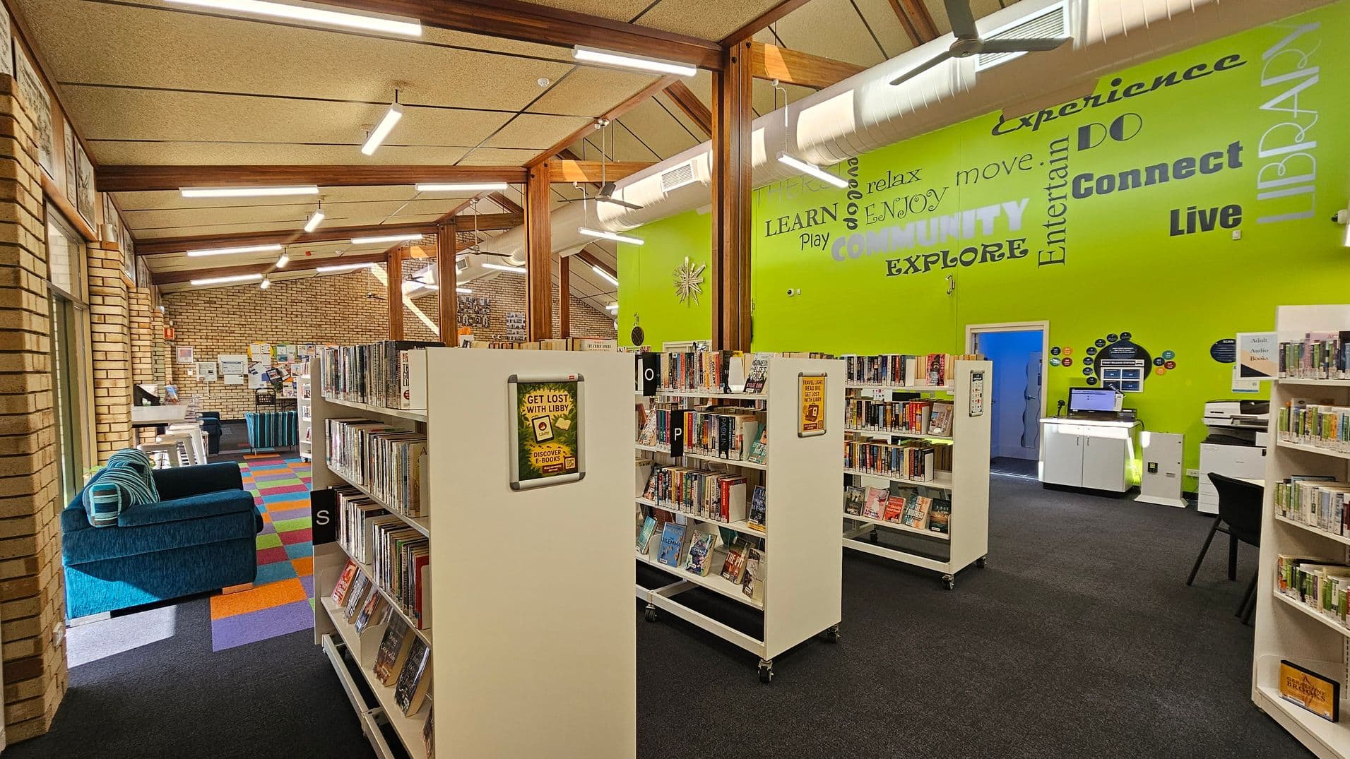 A library interior with bookshelves in the centre filled with books. The left side has a blue sofa and colourful carpet tiles. The right wall is bright green with words like "Experience," "Community," and "Explore" in various fonts. A computer station and printer are visible against the wall. The ceiling has exposed beams and fluorescent lights, and the floor is carpeted in dark grey.