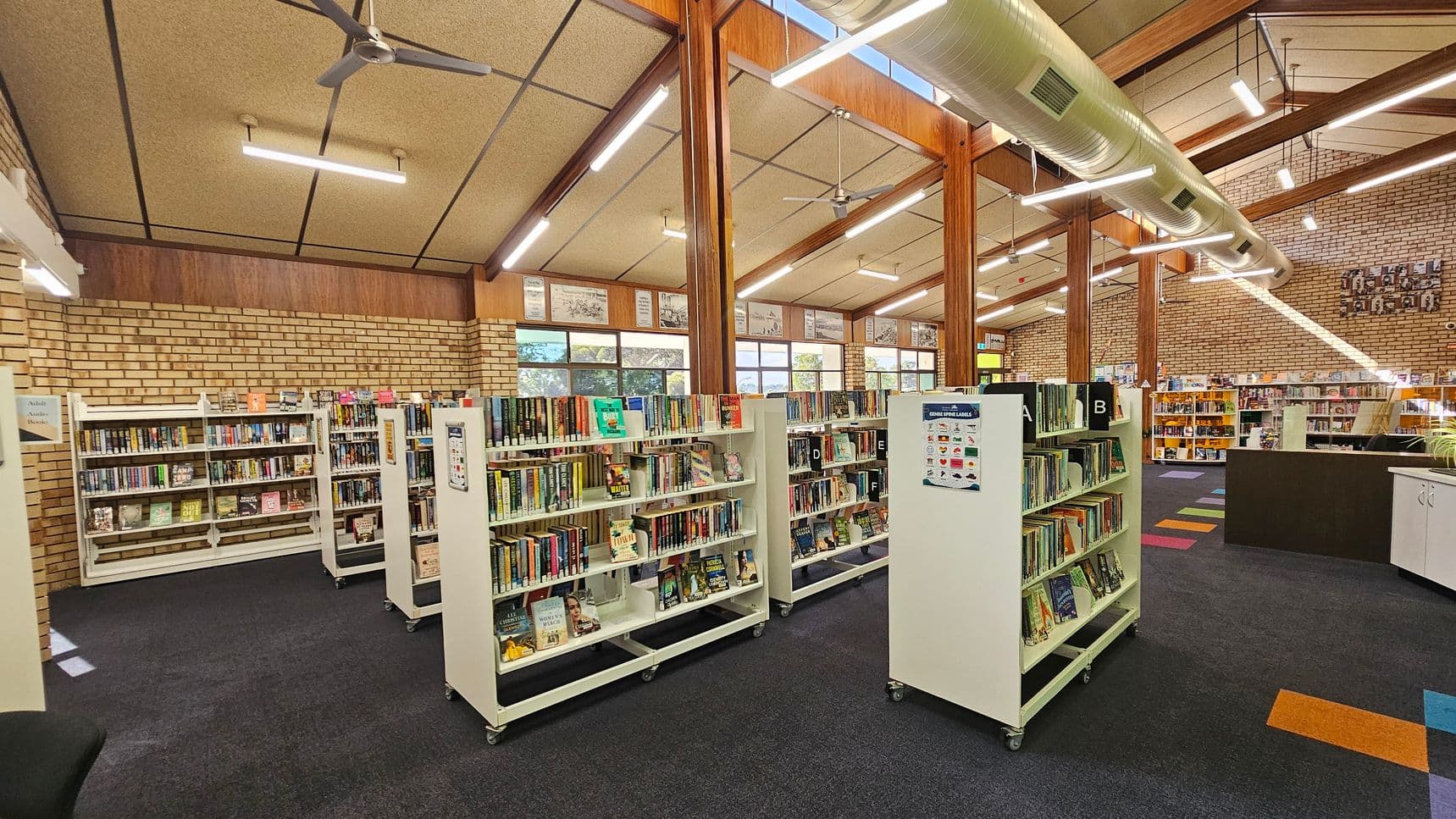 Bookshelves filled with books are arranged in rows on a dark carpeted floor. The ceiling is high with exposed beams, fluorescent lights, and ceiling fans. Large windows line the brick walls, allowing natural light to enter. A large air duct runs along the ceiling. In the background, more bookshelves and a counter are visible, with colourful carpet tiles scattered on the floor.