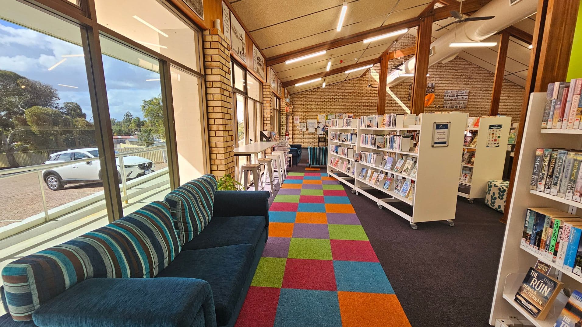 A library interior with a colourful, checkered carpet in the centre. On the left, large windows reveal a parked white car outside. A striped sofa is positioned along the windows. To the right, white bookshelves are filled with books. A high table with stools is near the windows. The ceiling is slanted with exposed beams and fluorescent lights. The walls are brick, and there are various notices and posters displayed.