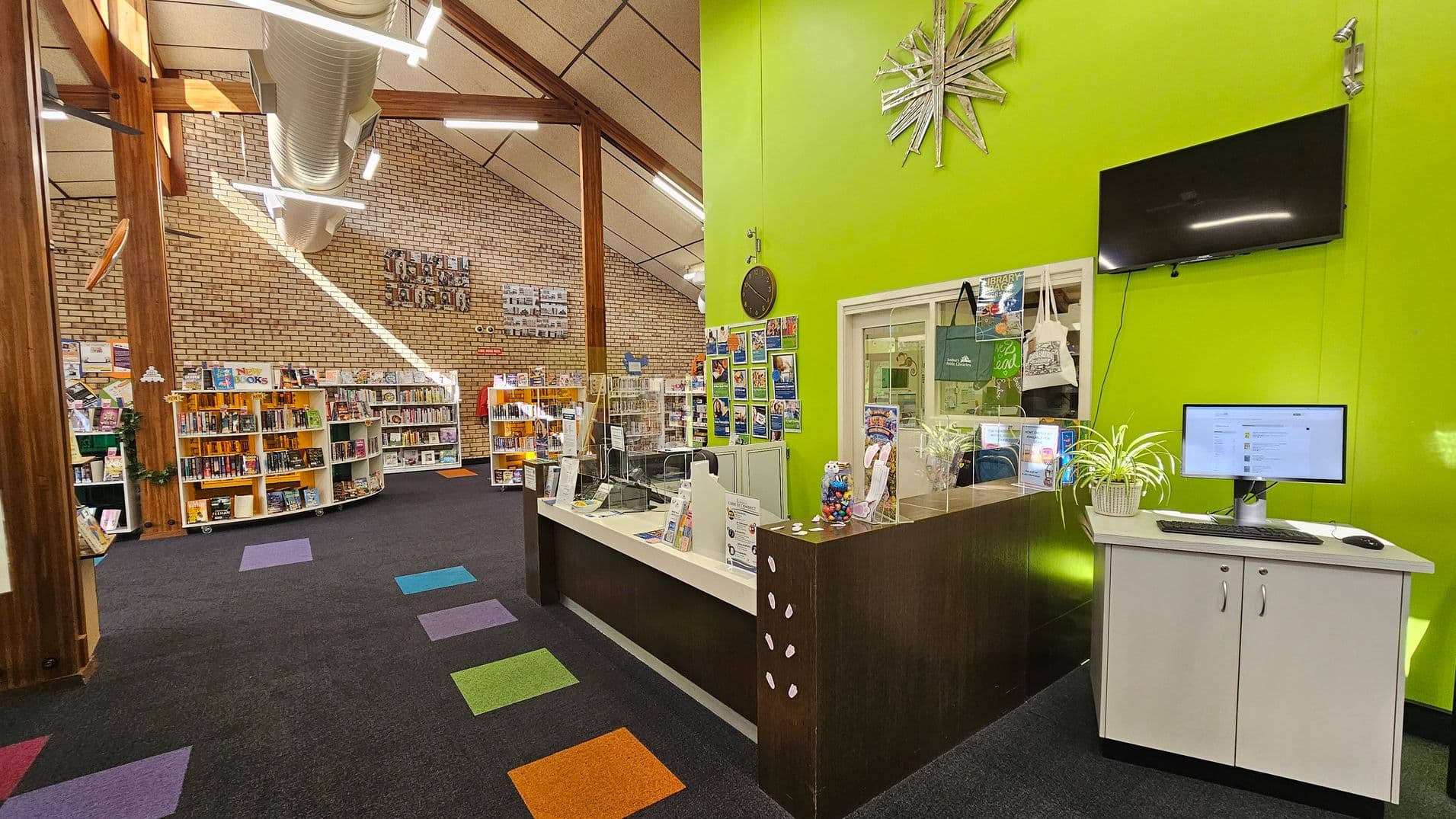 A library interior with a vibrant green wall on the right, featuring a clock, a large TV screen, and a computer on a white cabinet. The reception desk is adorned with informational pamphlets and a small plant. On the left, there are wooden beams and a brick wall with bookshelves filled with books. The ceiling is high with exposed ductwork and modern lighting. The carpet is dark with colourful square patterns scattered throughout.