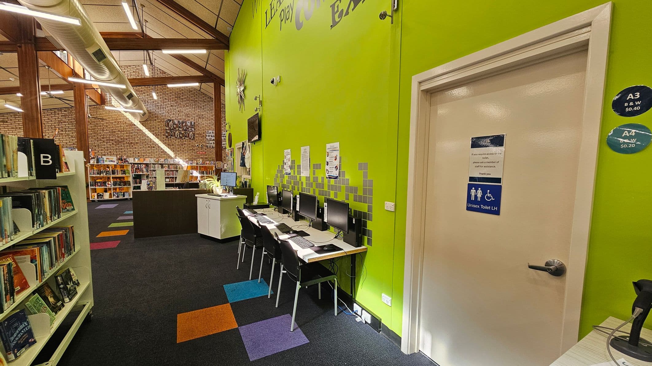 A library interior with a bright green wall on the right featuring a row of computers and chairs. Above, there are words partially visible, such as "Learn" and "Explore." A door marked "Unisex Toilet LH" is also on the right. On the left, bookshelves labelled with letters and filled with books. The floor is carpeted with colourful square patterns. The ceiling is high with exposed beams and ductwork, and the back wall is made of brick with more bookshelves and a counter.