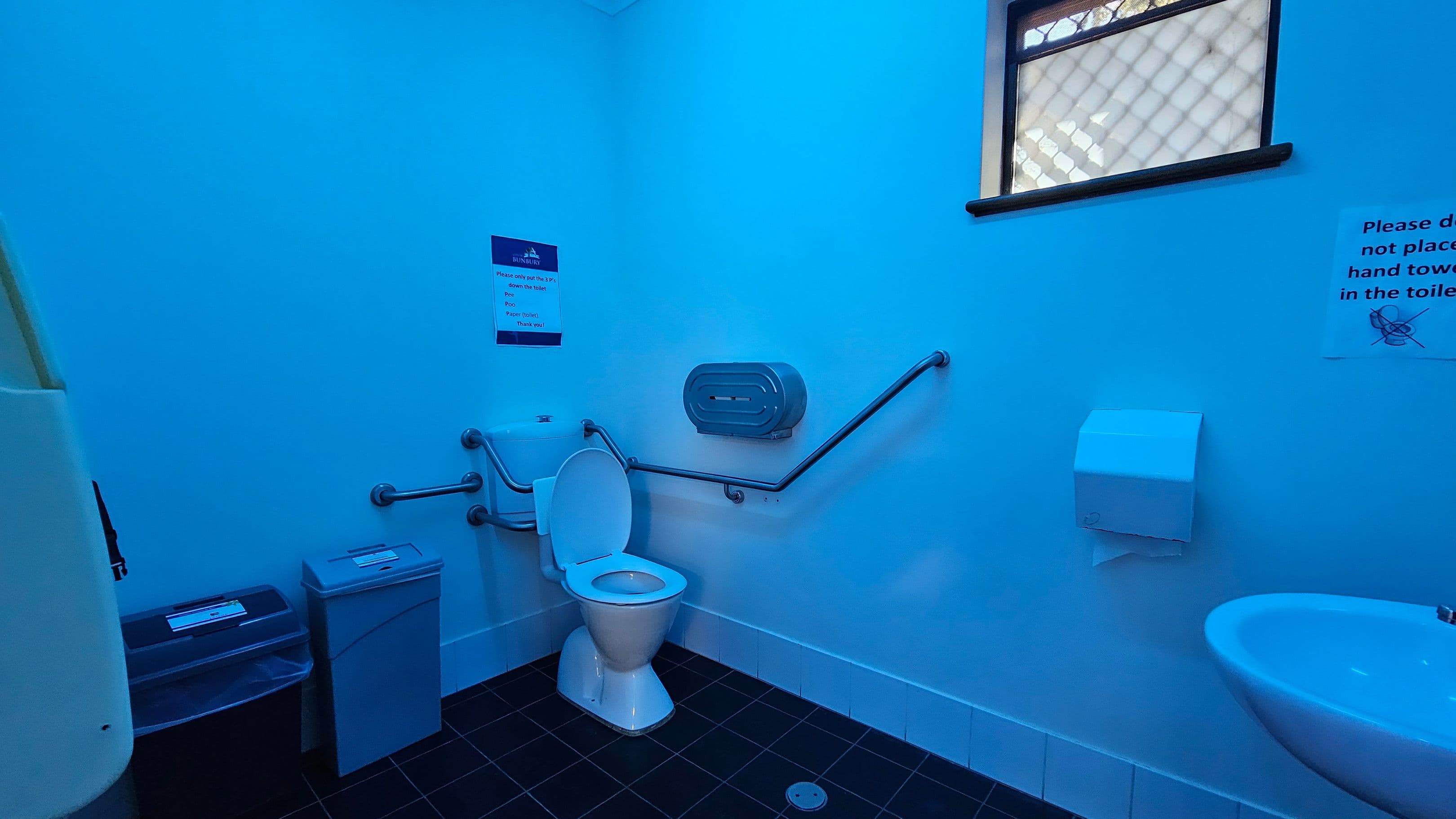 A bathroom with blue lighting. On the left, there are two bins, one with a black lid and the other with a grey lid. In the centre, a white toilet with grab bars on either side is mounted against the wall. Above the toilet, a sign is visible. To the right, a toilet paper dispenser and a paper towel dispenser are mounted on the wall. A small sink is positioned below the paper towel dispenser. A window with a mesh covering is on the upper right wall. The floor is tiled in dark grey.
