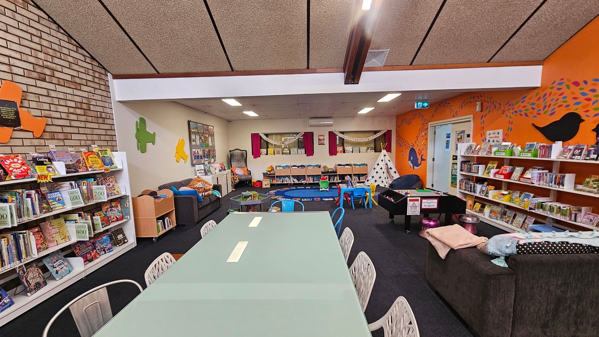 A colourful children's library area with a long table and chairs in the foreground. To the left, bookshelves are filled with various books, and a brick wall features colourful plane cutouts. In the centre, a cosy seating area with sofas and cushions is visible, along with a play area featuring a small tent and toys. The right side has more bookshelves against an orange wall decorated with bird silhouettes. The ceiling is lined with panels and fluorescent lights.